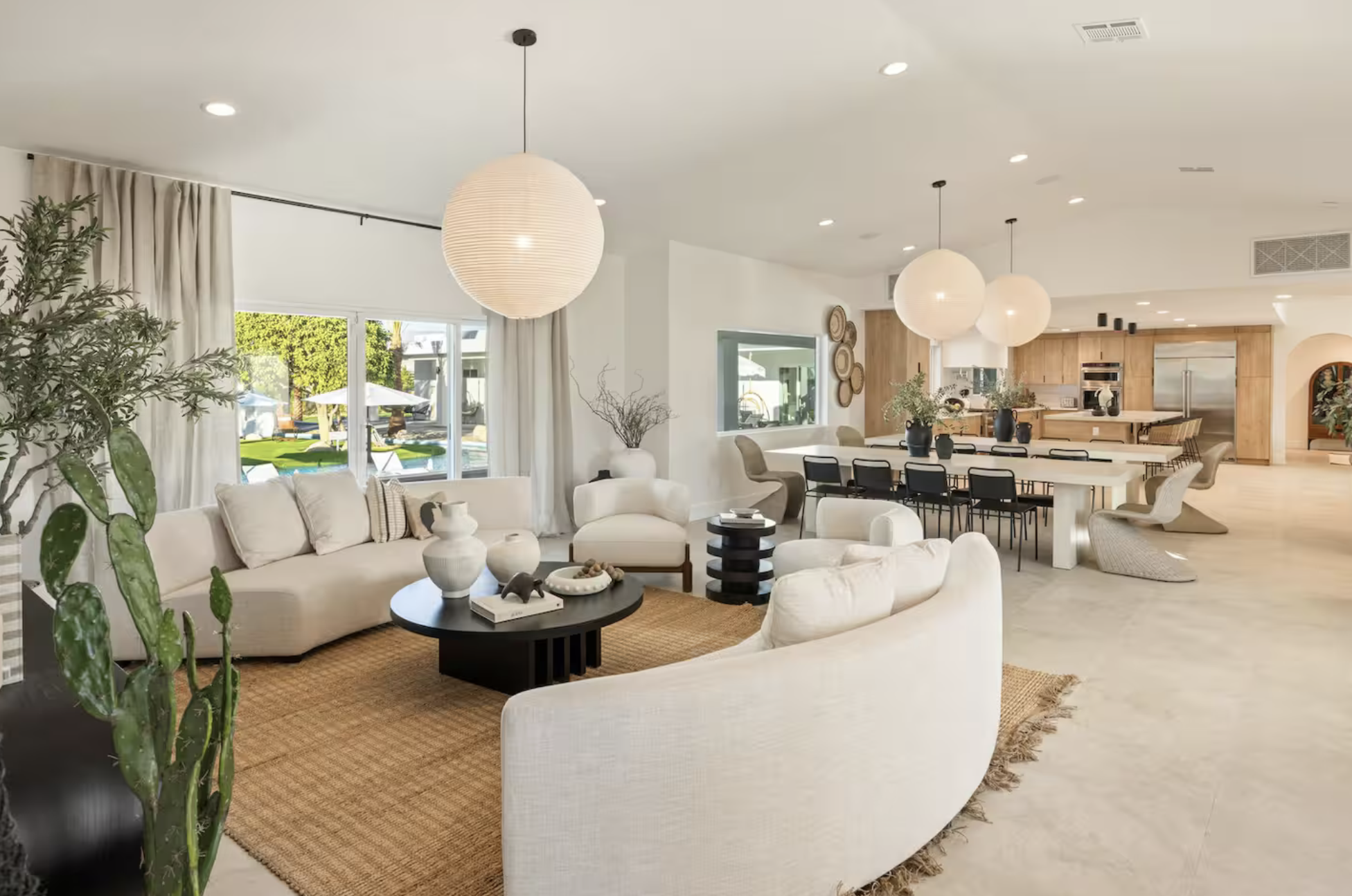 Open-concept living and dining area with white walls, wooden accents, round hanging lights, large windows, and outdoor view