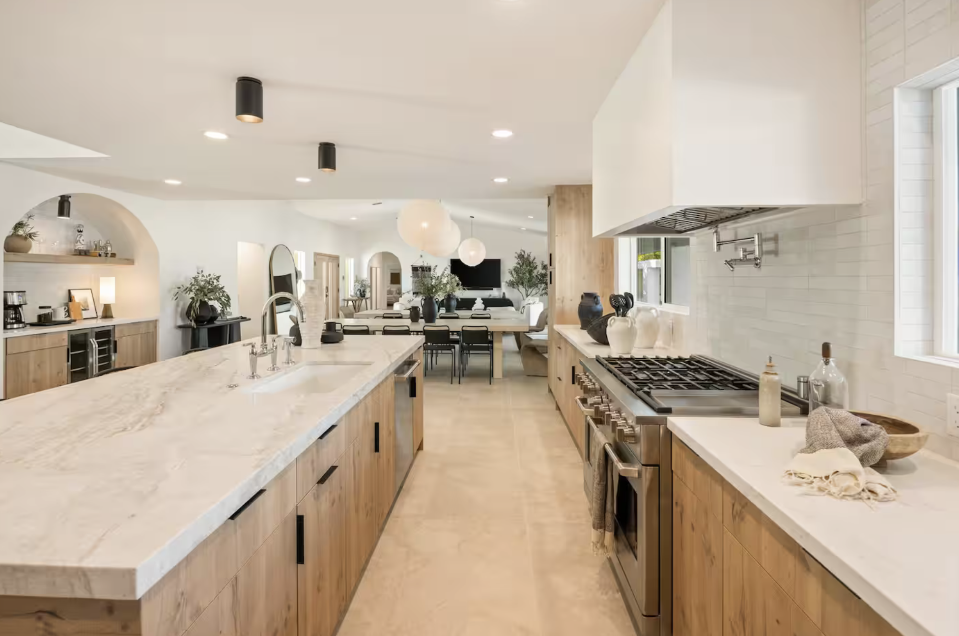 Modern open-concept kitchen with wooden cabinetry, marble countertops, black appliances, and a spacious dining area with white and black furniture, decorated with plants and pendant lights.