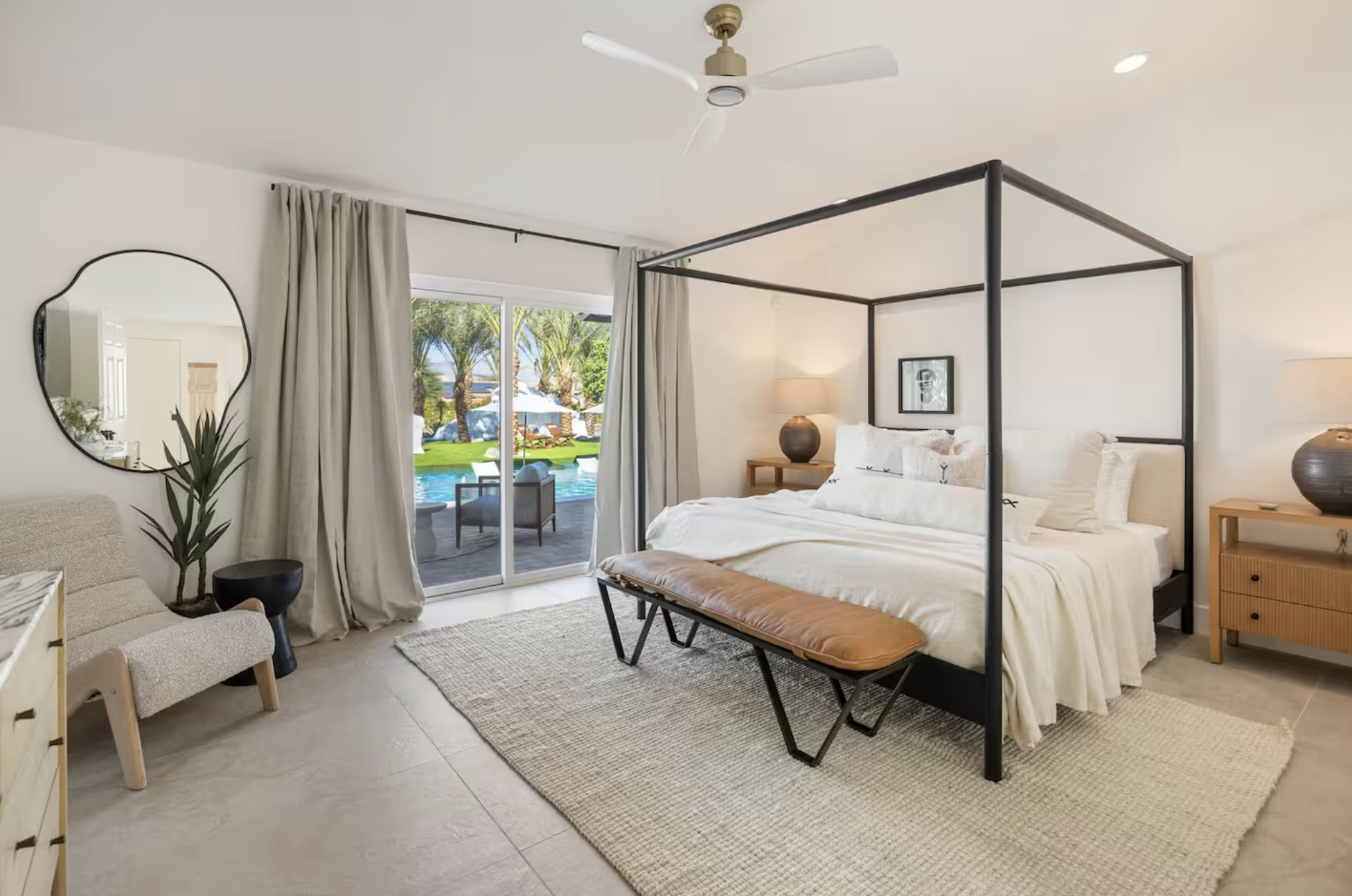 A modern bedroom with a black canopy bed, bedside tables with lamps, a bench, and a sliding glass door leading to a backyard with a pool and lounge chairs.