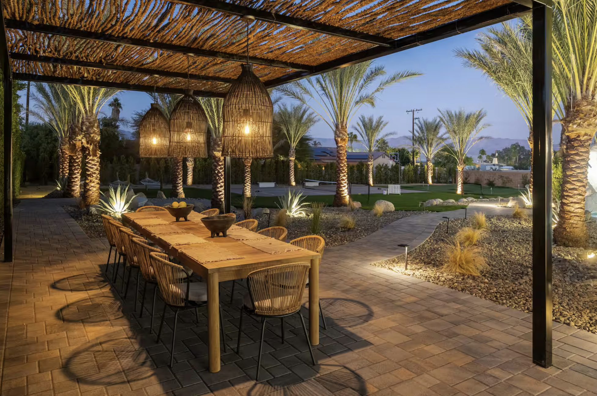 Outdoor patio area with a large wooden dining table, wicker chairs, hanging wicker pendant lights, palm trees, landscaped yard, and pathway with landscape lighting at dusk.
