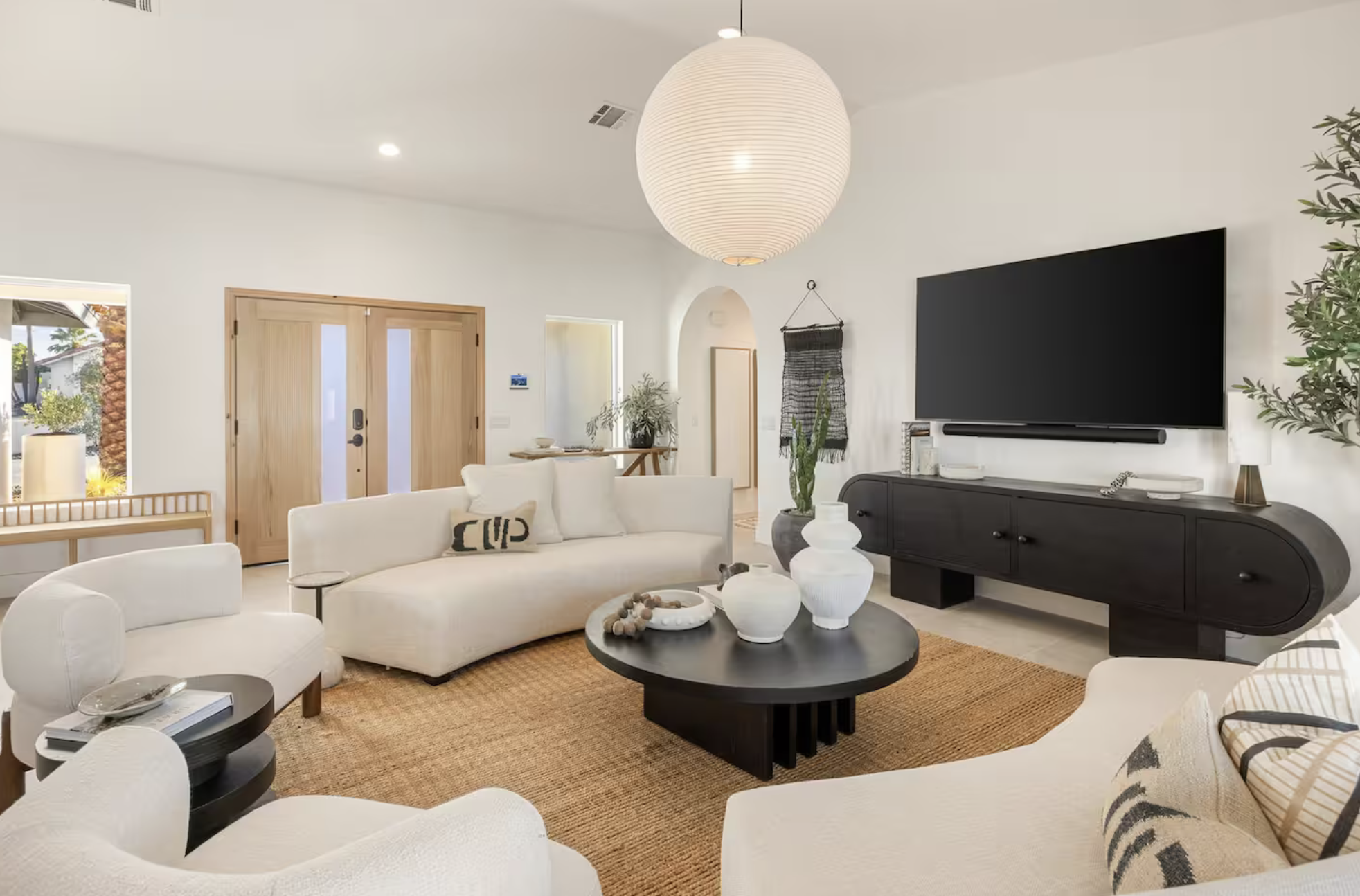 Living room with white walls, large white sofa, white armchair, black coffee table with white vases, black TV stand with a large flat-screen TV, potted plants, woven rug, and wooden front door, illuminated by natural and ceiling lighting.