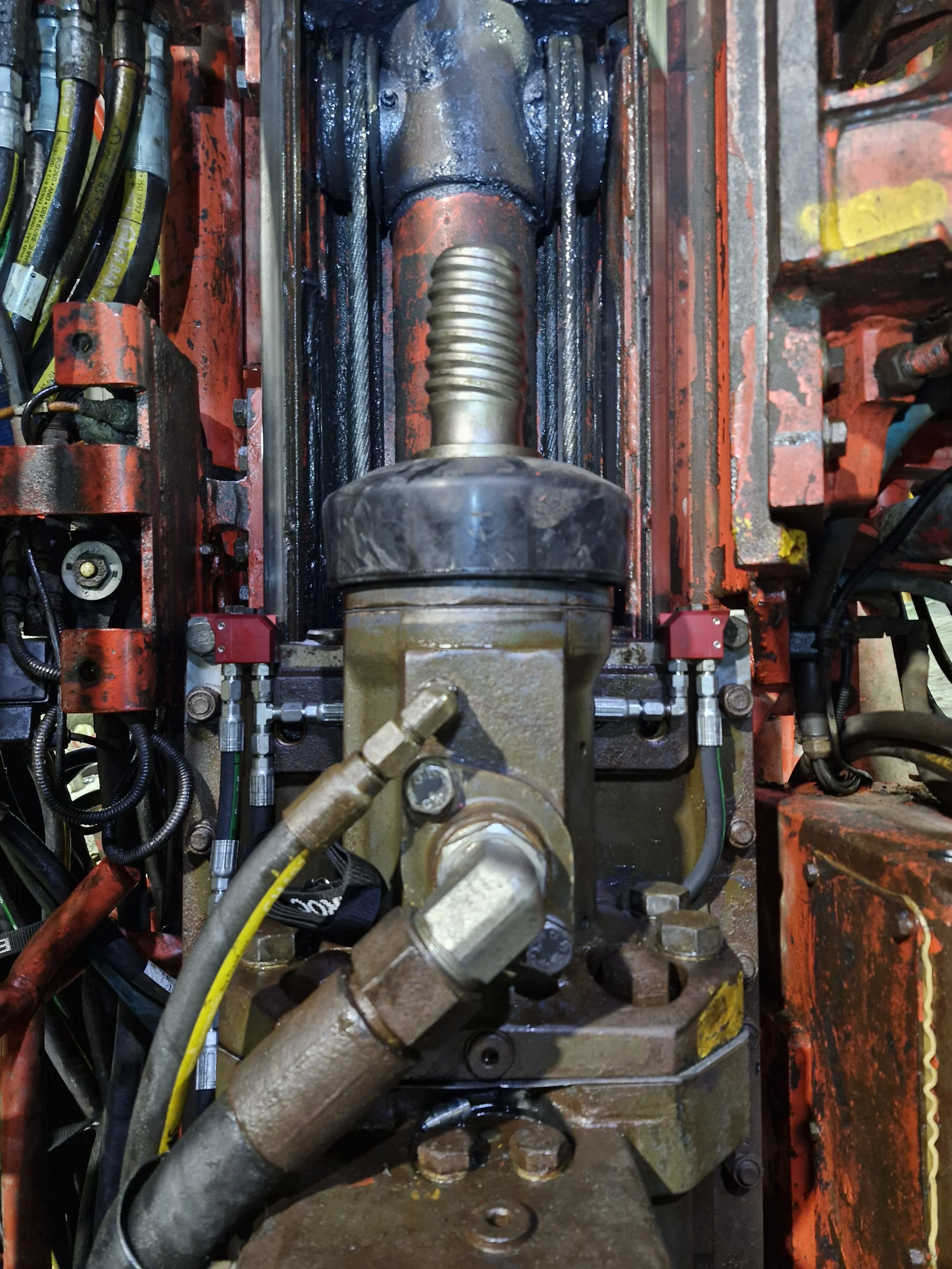 A section of a complex hydraulic or mechanical system with various pipes, hoses, and metallic components, including a prominent large bolt and some wiring on the sides, with some parts enclosed in a red frame.