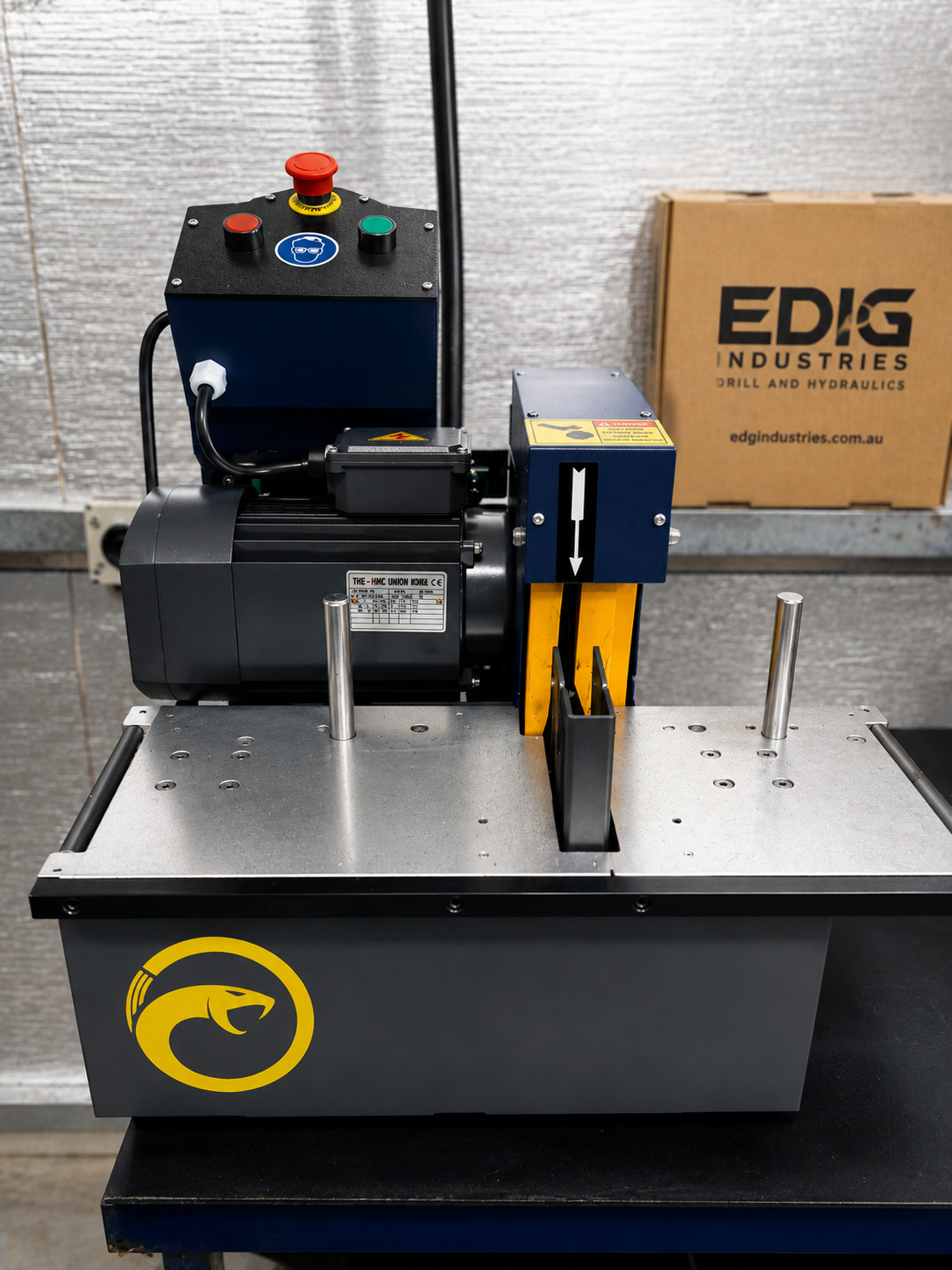 A hydraulic cutting machine with a control panel, mounted on a table, and a yellow and black drilling slot, near a cardboard box labeled 'EDG Industries'.