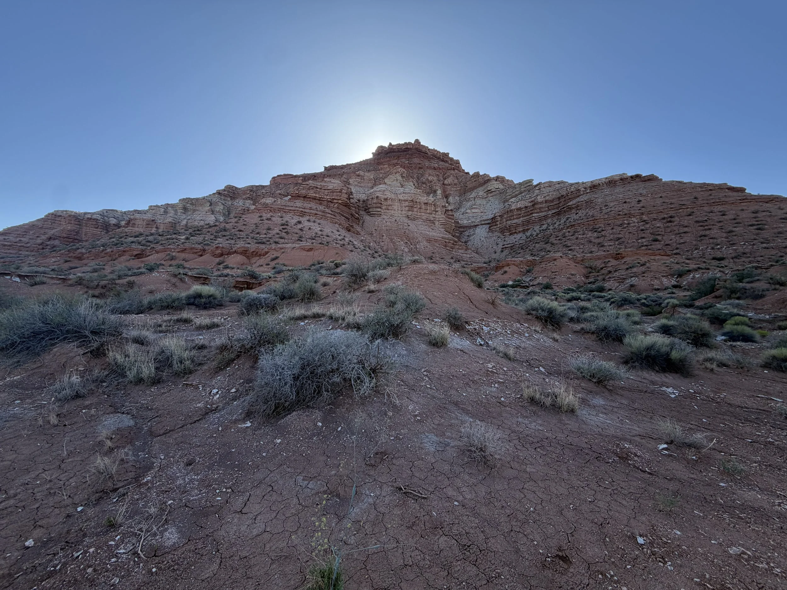 A dry, cracked desert landscape with sparse vegetation and layered red and white rock formations under a clear blue sky with the sun behind the rocks.