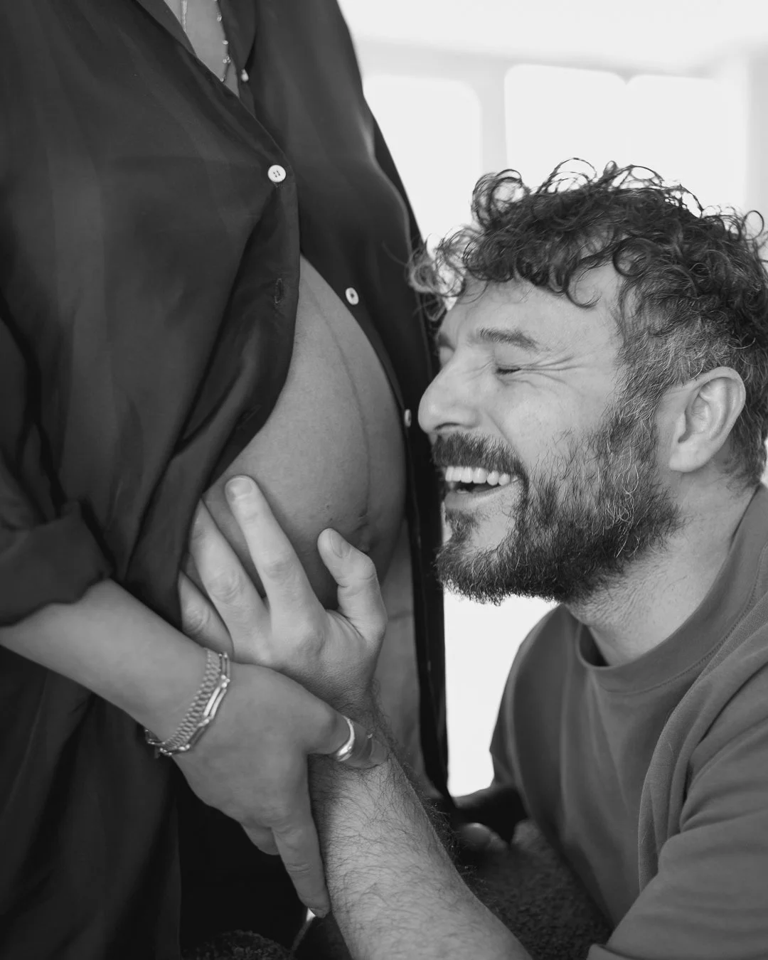 A man with curly hair and a beard happily looking at a pregnant woman, gently touching her belly, in a black and white photo.