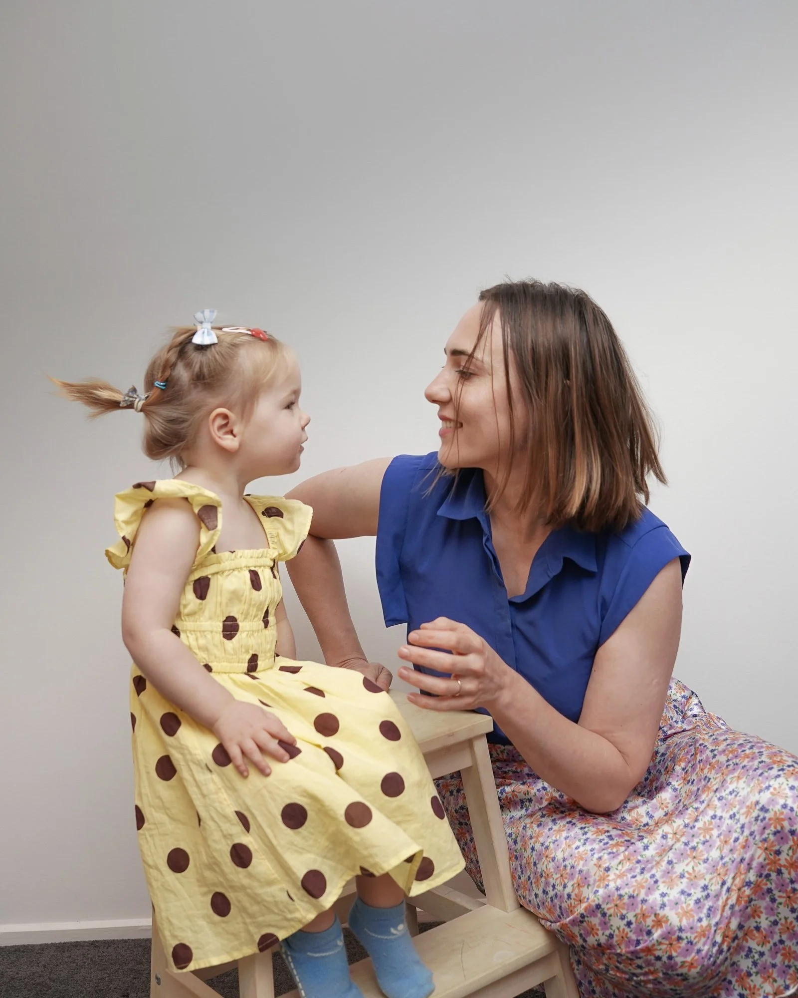 A young girl in a yellow dress with brown polka dots sitting on a wooden chair, facing a woman in a blue shirt and floral skirt, who is smiling at her and leaning toward her, in a room with plain gray walls.