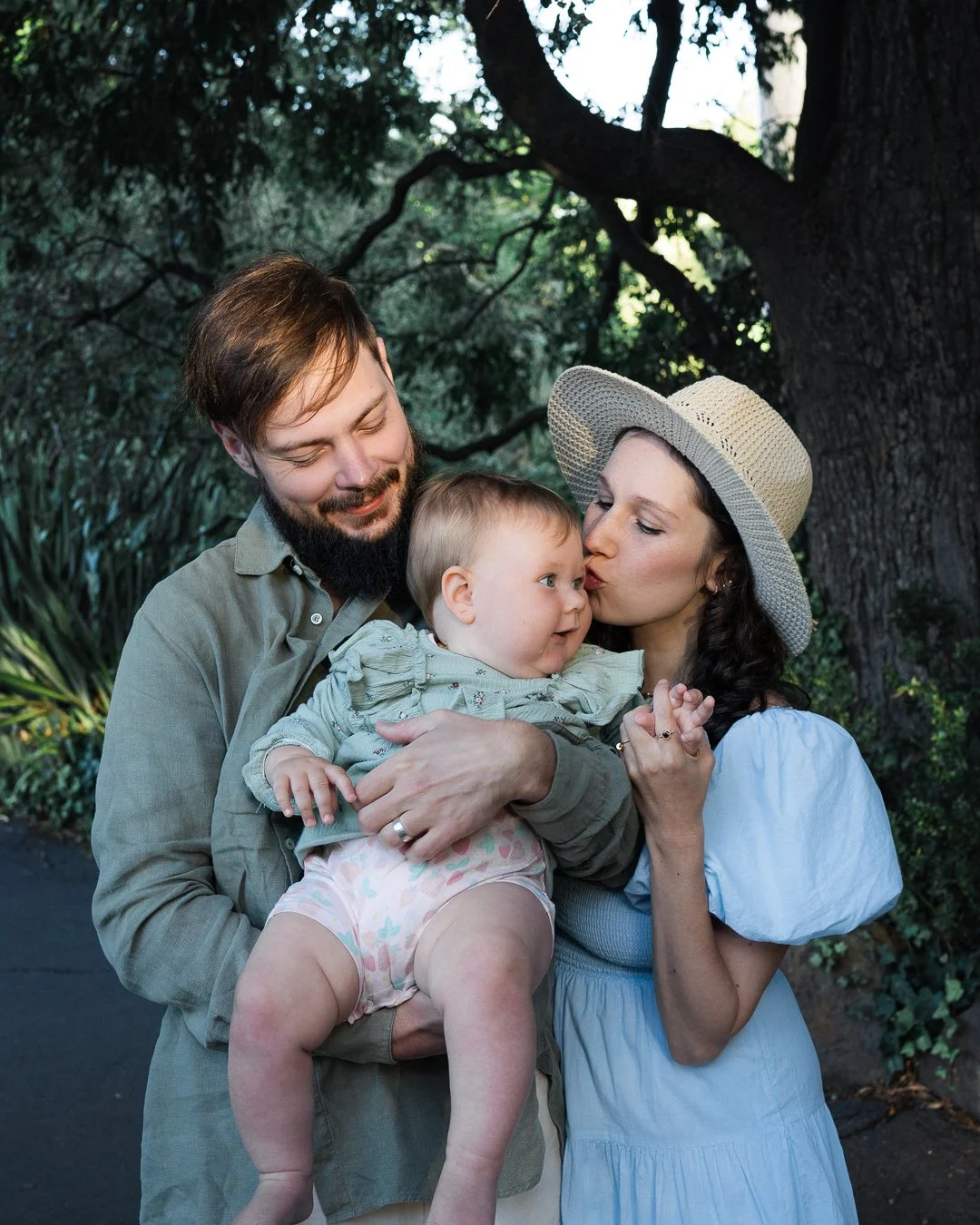 Parents cuddling and kissing their baby during an outdoor family photography session at the Royal Botanic Gardens in Melbourne.