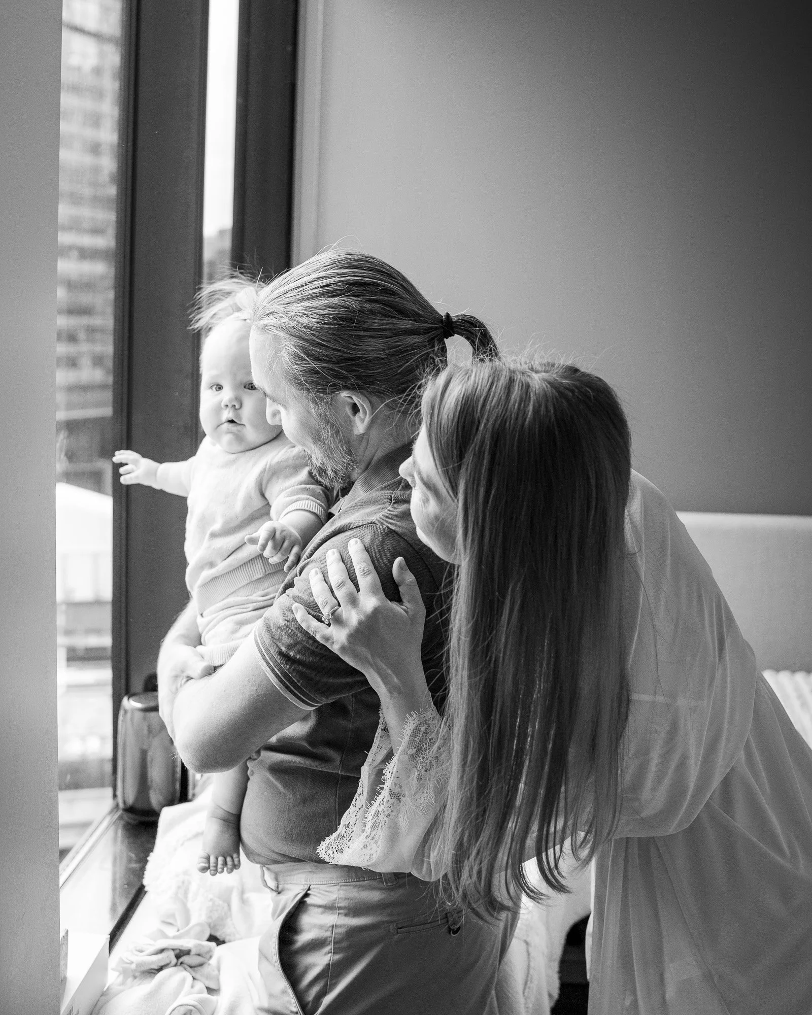 Parents cuddling their baby by a window during an in-home family photography session in Melbourne, while the baby looks curiously outside.