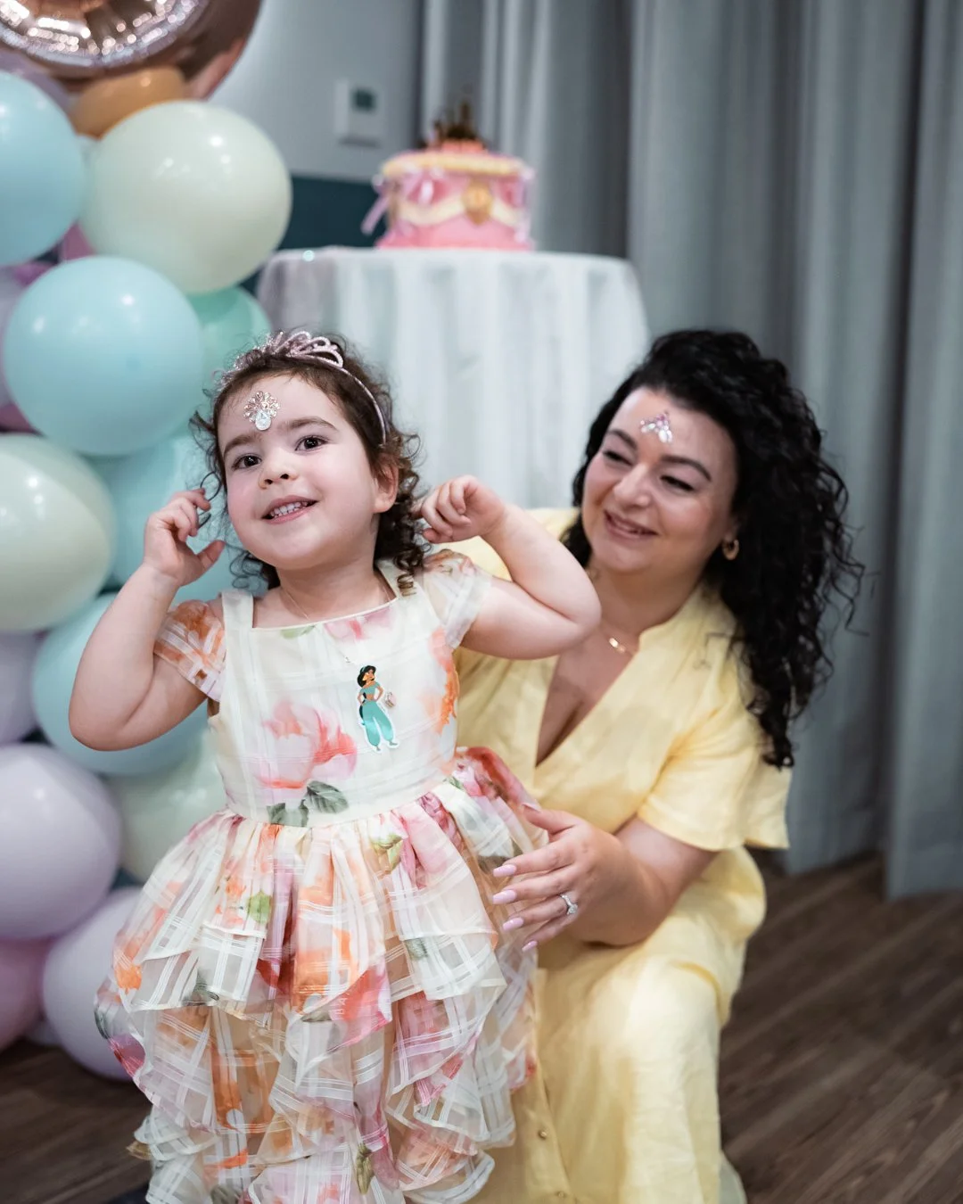 A little girl smiling while celebrating her birthday with her mother beside pastel balloons and a birthday cake during a family celebration in Melbourne.