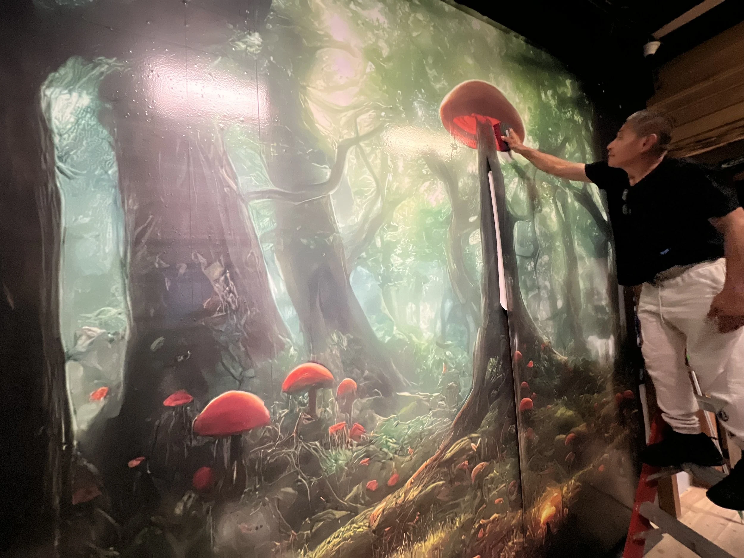 A person standing next to a large vibrant mural of a forest scene with red mushrooms and green foliage, reaching out with their right hand.