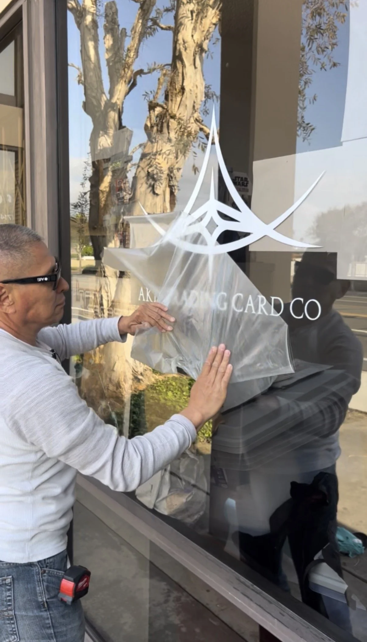 Two men are applying a frosted sticker with a geometric design and the words 'AKADIN GARD CO' onto a glass window outside a building. One man is holding the sticker while the other smooths it out. The background shows a large tree and a sunny day outside.
