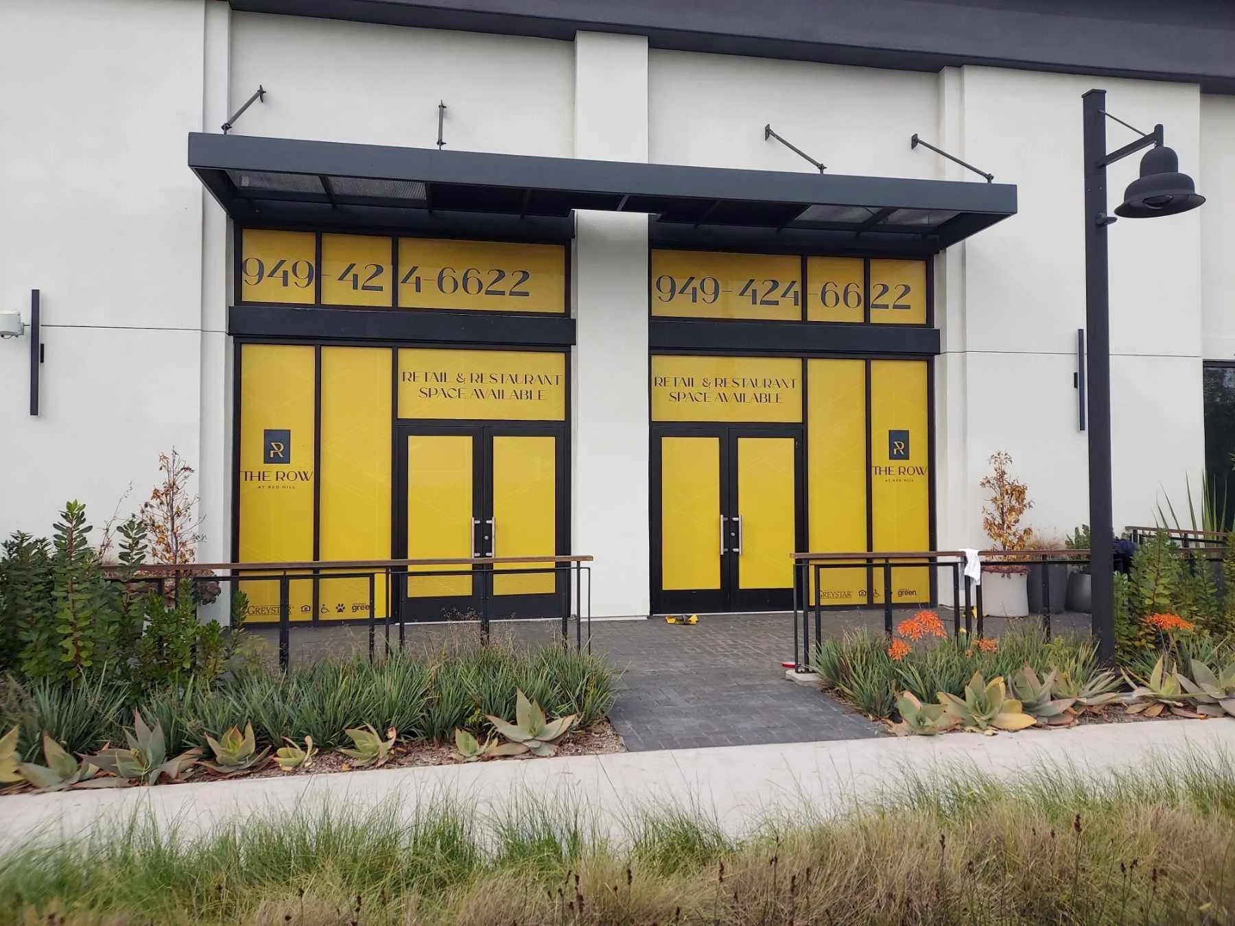 Storefront with yellow doors and black frames displaying the phone number 949-424-6622 and the text 'RETAIL & RESTAURANT SPACE AVAILABLE,' surrounded by plants and a sidewalk, with black street lamps and a black awning above.