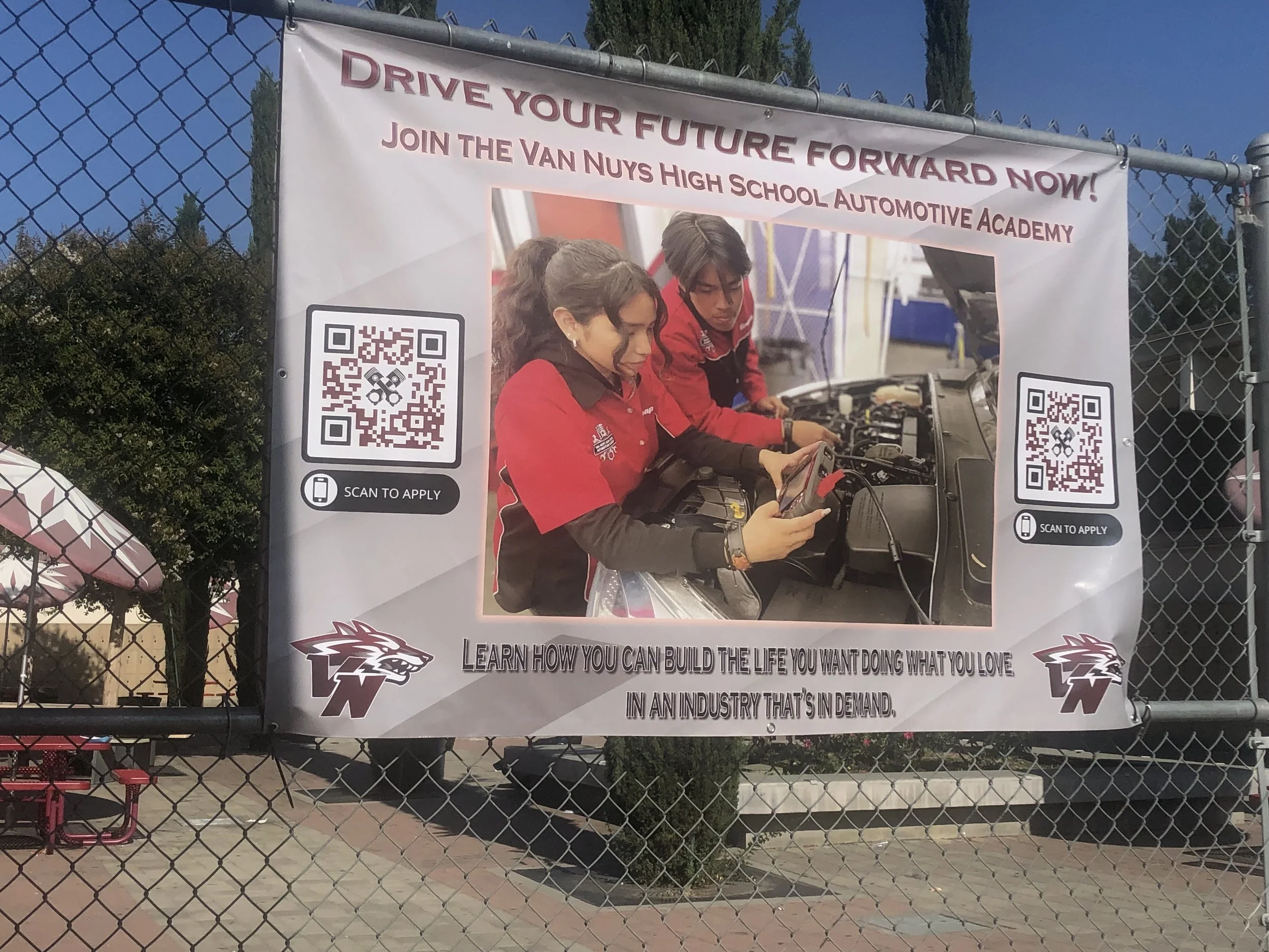 A banner hanging on a chain-link fence promoting Van Nuys High School Automotive Academy, with students working on a car engine in the background and QR codes for application. The banner has text saying, 'Drive Your Future Forward Now! Join the Van Nuys High School Automotive Academy. Learn how you can build the life you want doing what you love in an industry that's in demand.'
