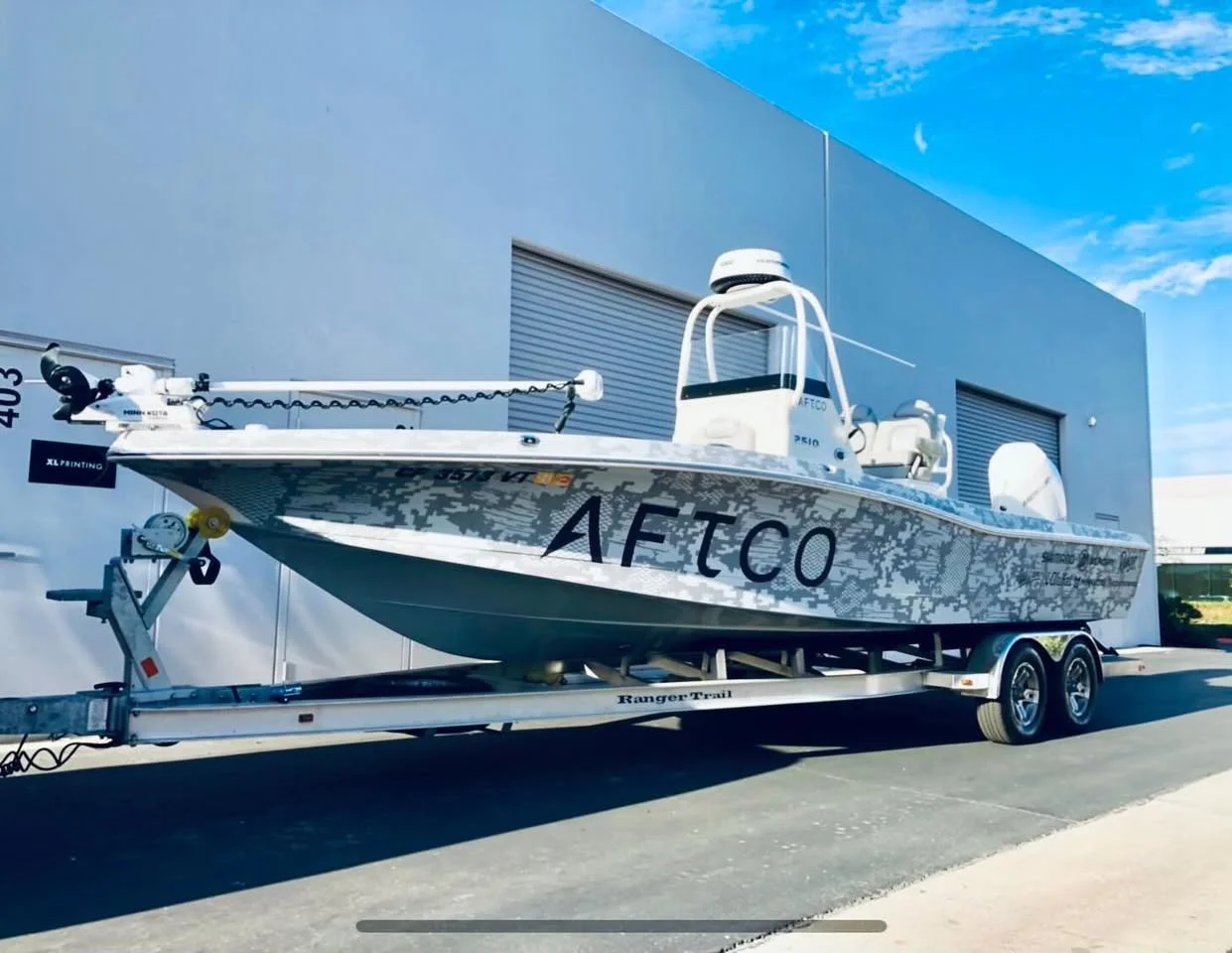 A white fishing boat with camouflage pattern on the side, named AFTO, on a trailer parked outside a building with blue sky overhead.