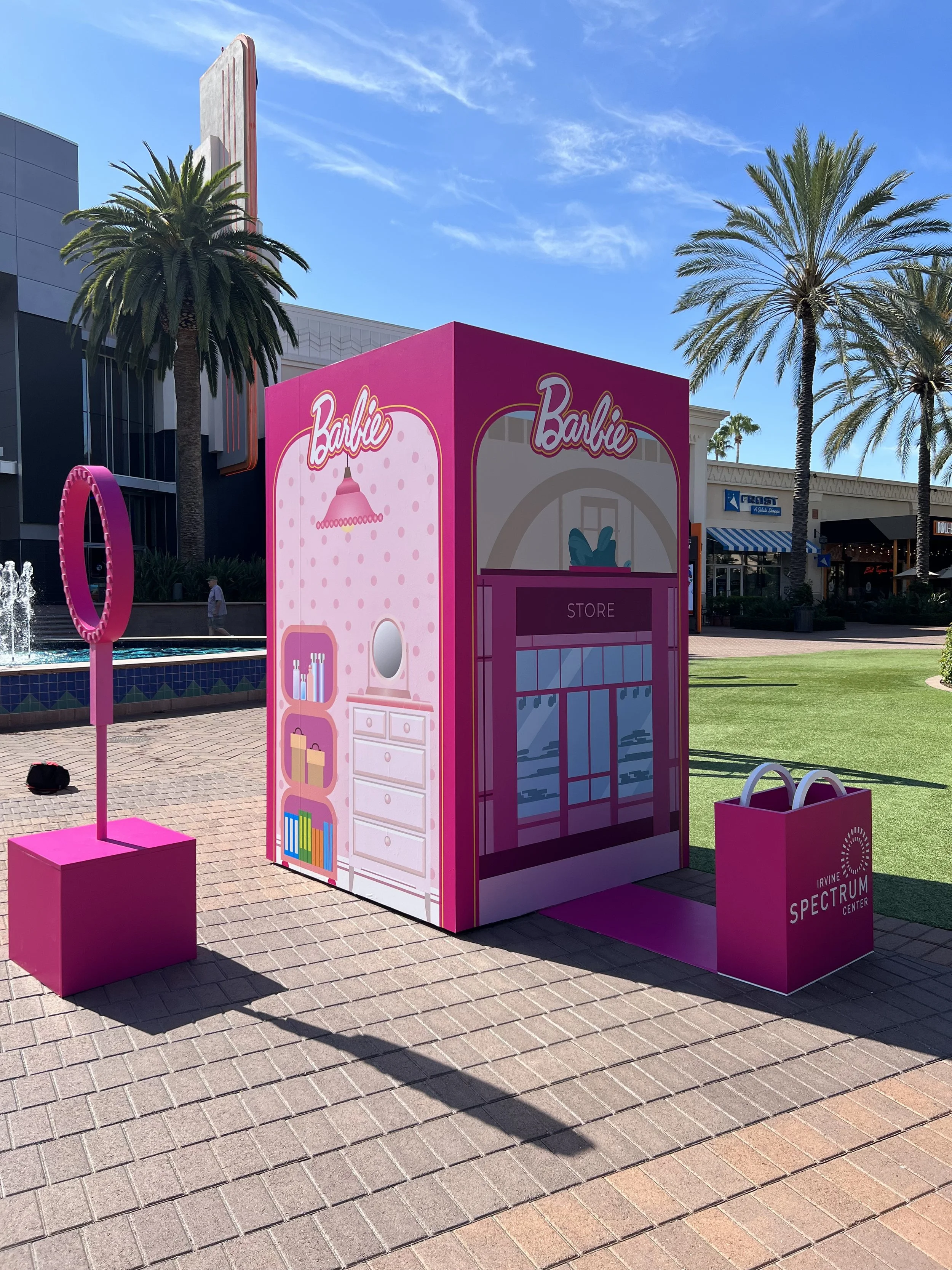 Pink promotional display for Barbie at Irvine Spectrum Center, with palm trees, a water fountain, and stores in the background on a sunny day.