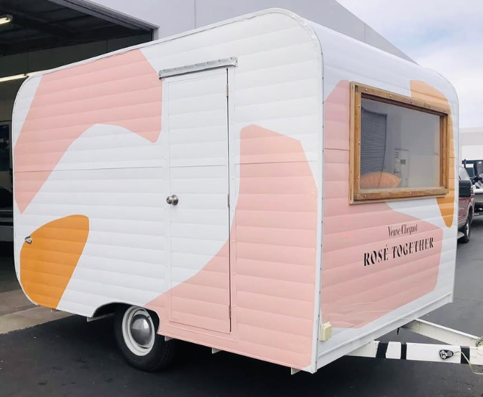 A small camping trailer painted with pink, white, and orange abstract shapes, featuring a wooden framed window and a door, with the text 'Venue Chapel ROSE TOGETHER' on the side.