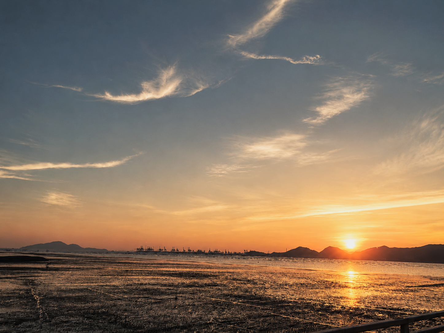 Sunset over a coastal landscape with mountains in the background, reflective water, and industrial structures on the horizon.