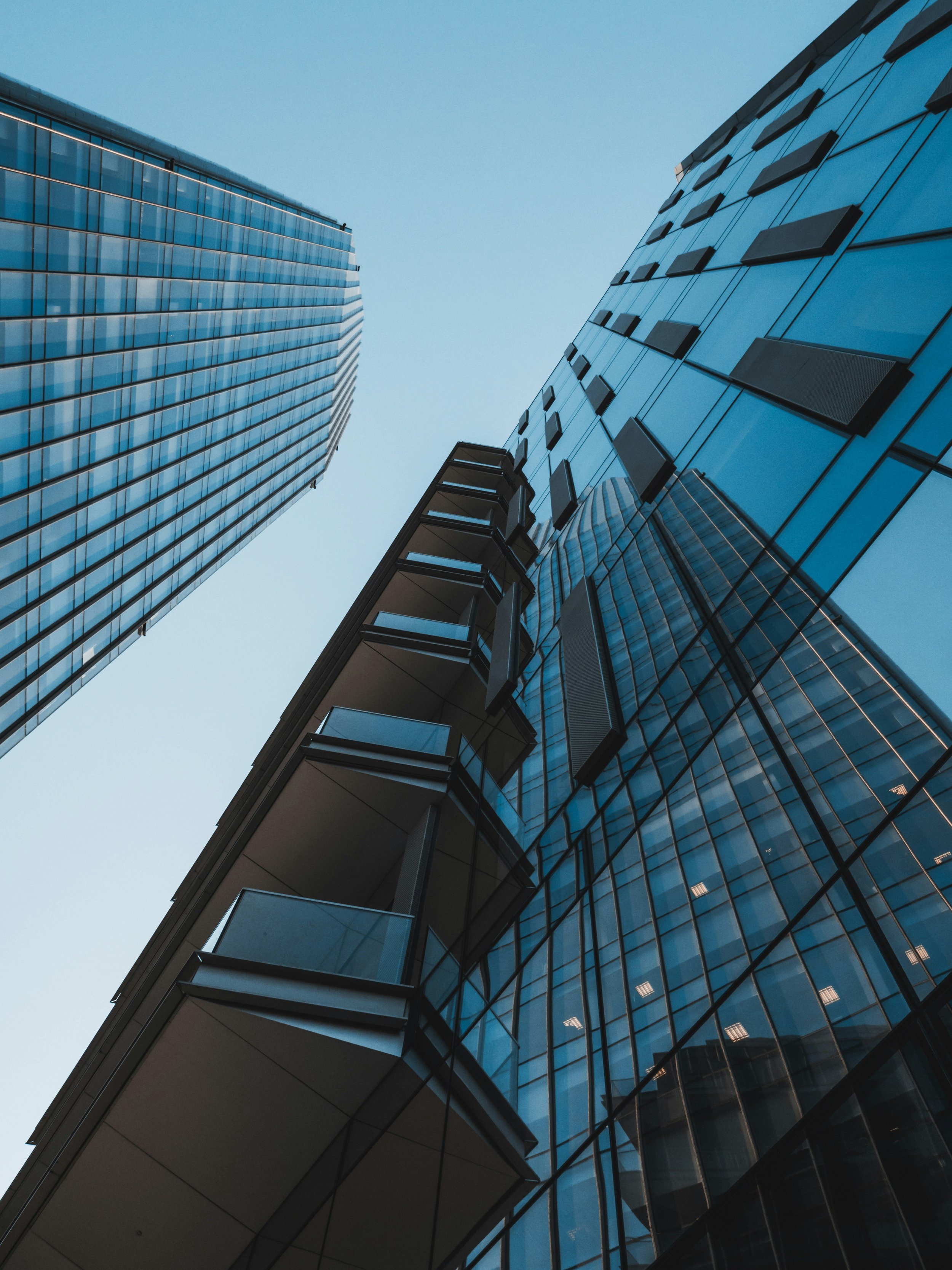 Looking up at tall modern glass skyscrapers from the ground, with reflections on the windows and a clear blue sky above.