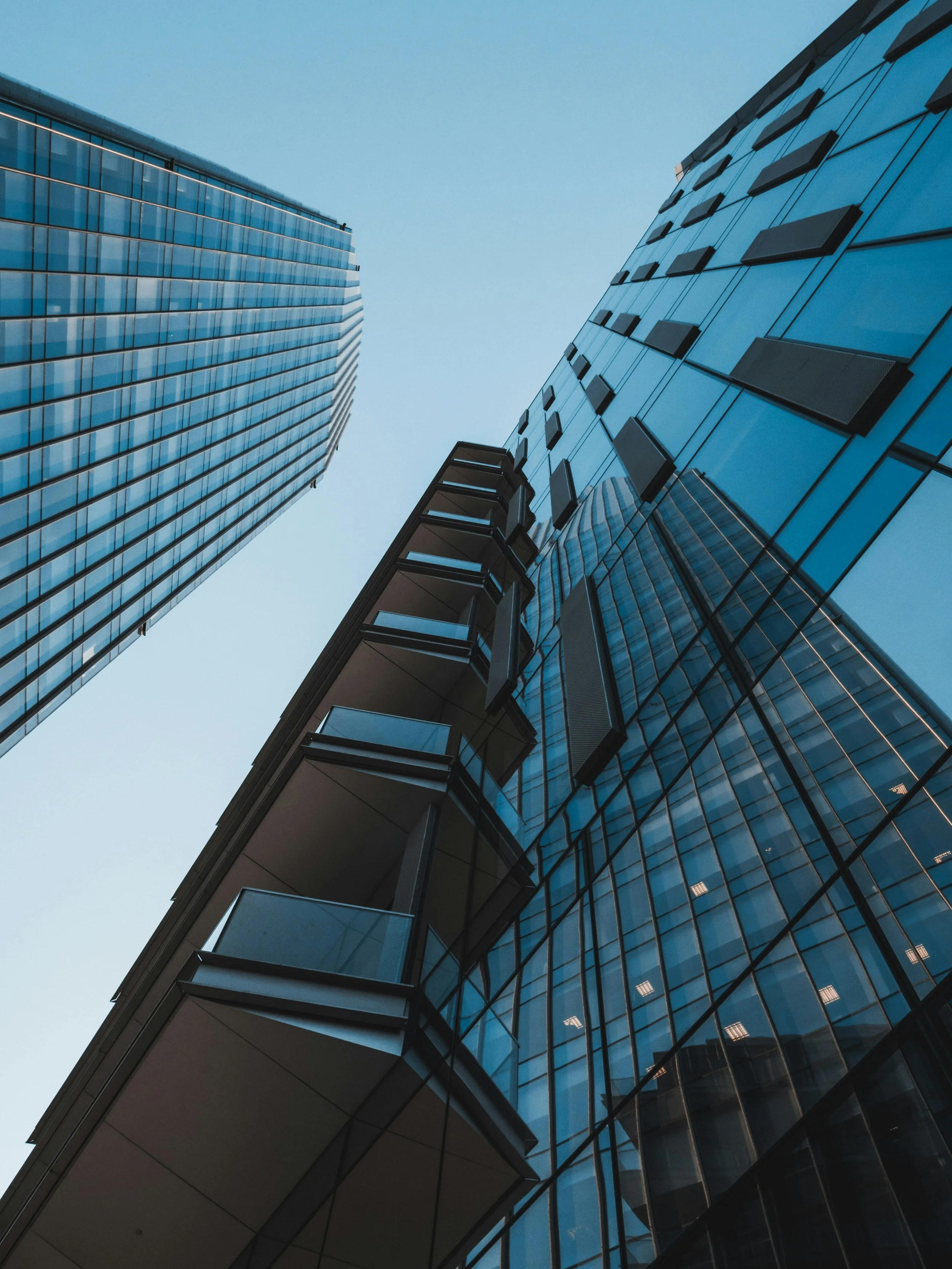 Looking up at modern glass skyscrapers with reflective windows and multiple balconies against a clear blue sky.