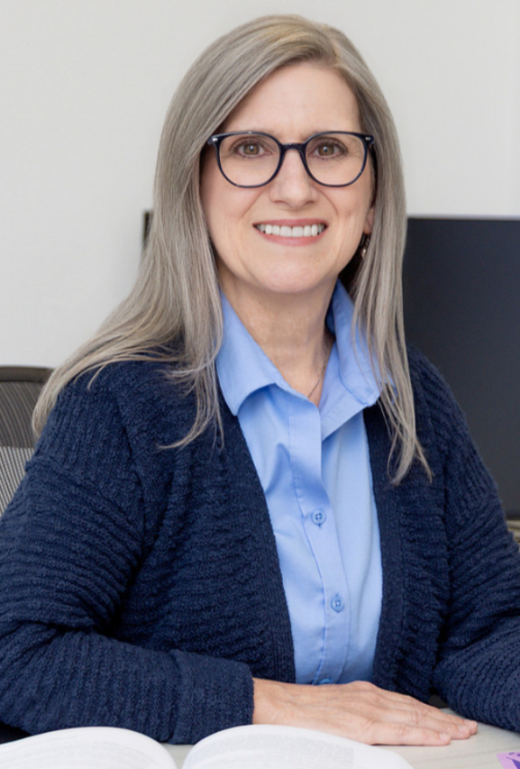 A woman with glasses and shoulder-length gray hair sitting at a desk in an office environment, smiling at the camera.