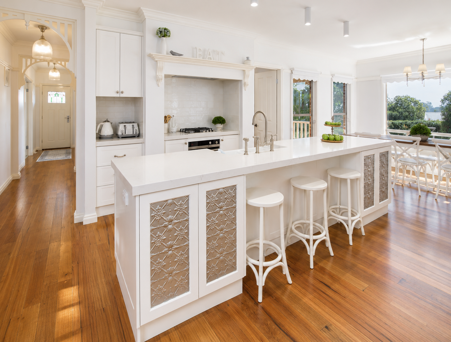 Bright, airy kitchen with white cabinets and countertops, wooden floors, and a large island with bar stools. There are three windows letting in natural light, a dining area with table and chairs, and decorative items like green apples and plants.