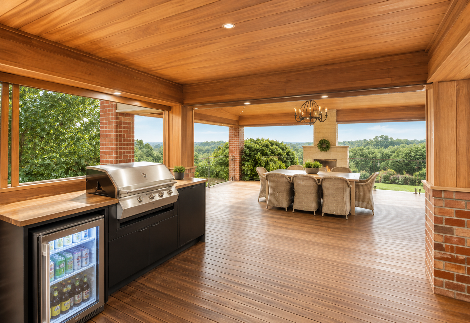 Covered outdoor patio with wooden ceiling and flooring, outdoor grill, indoor-style dining table with wicker chairs, fireplace, chandelier, and a view of lush green trees and landscape.