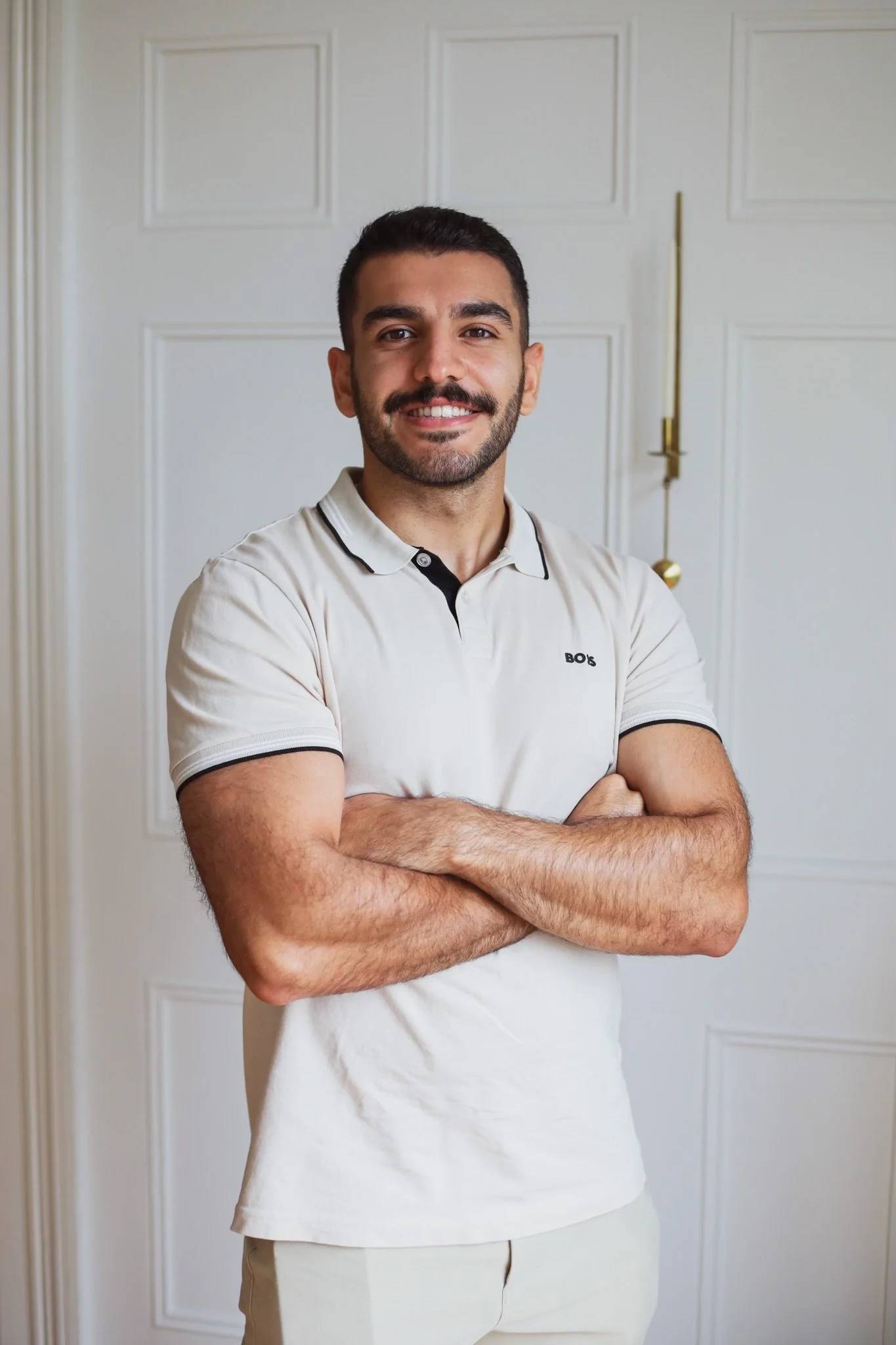 A smiling man with dark hair and a beard standing with his arms crossed in front of a white paneled door.
