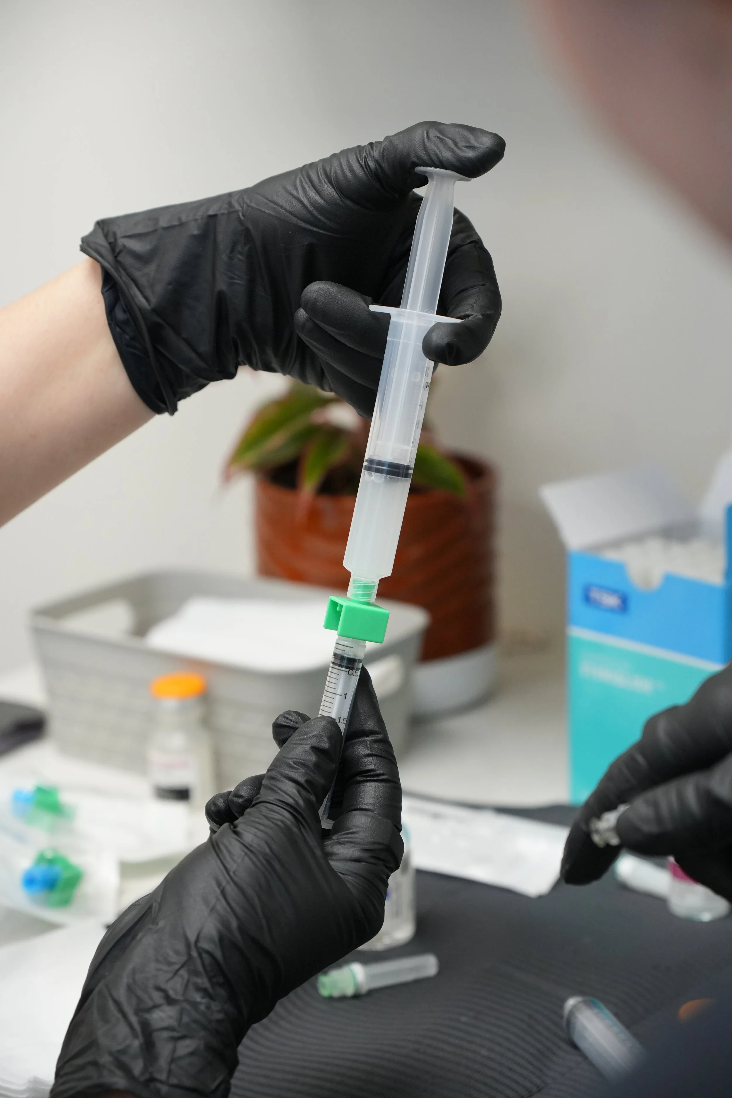 A person wearing black gloves holding a syringe in a laboratory setting. There are various vials and medication items on a white surface in the background.