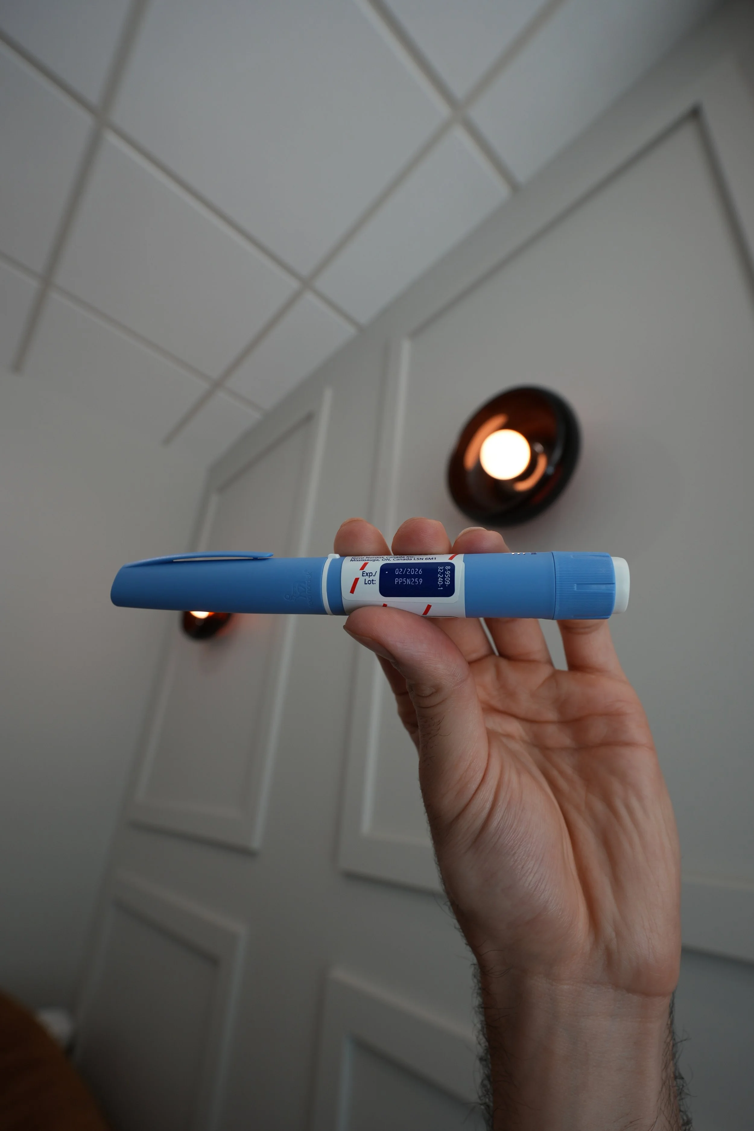 A person holding a blue and white medical test tube or sample container with a label, with a ceiling and a wall with white paneling and a ceiling vent in the background.