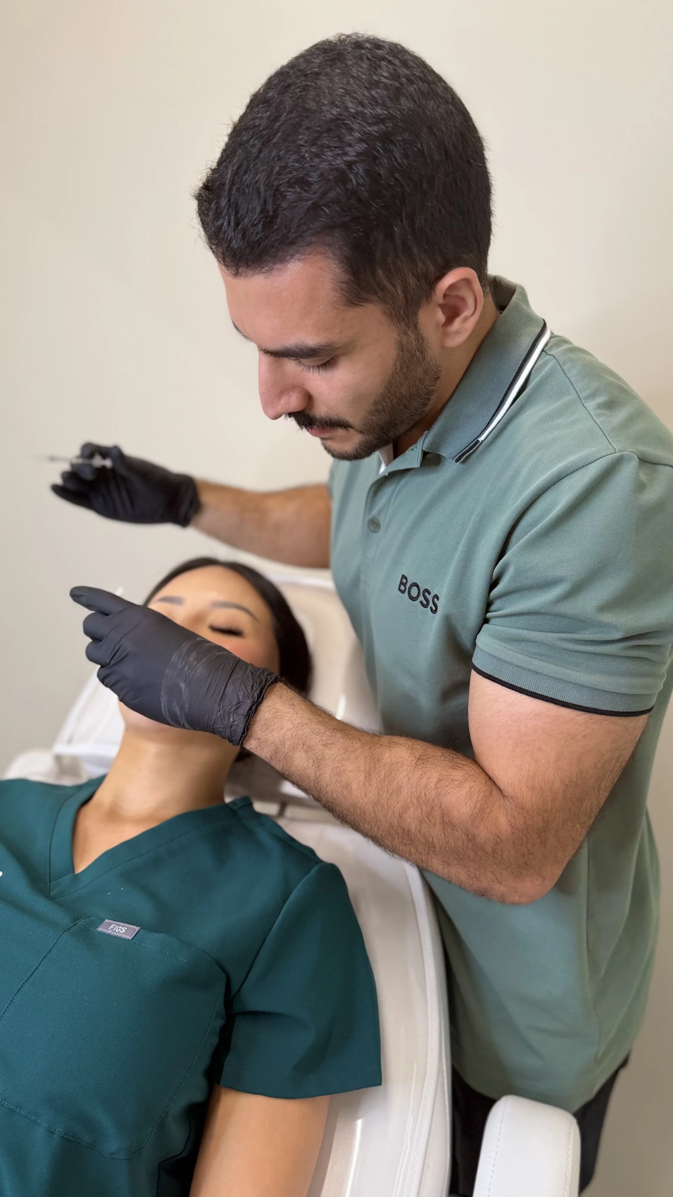 A man in a green shirt labeled 'BOSS' administers an injection to a woman lying on a medical bed, with her eyes closed, in a clinical setting.
