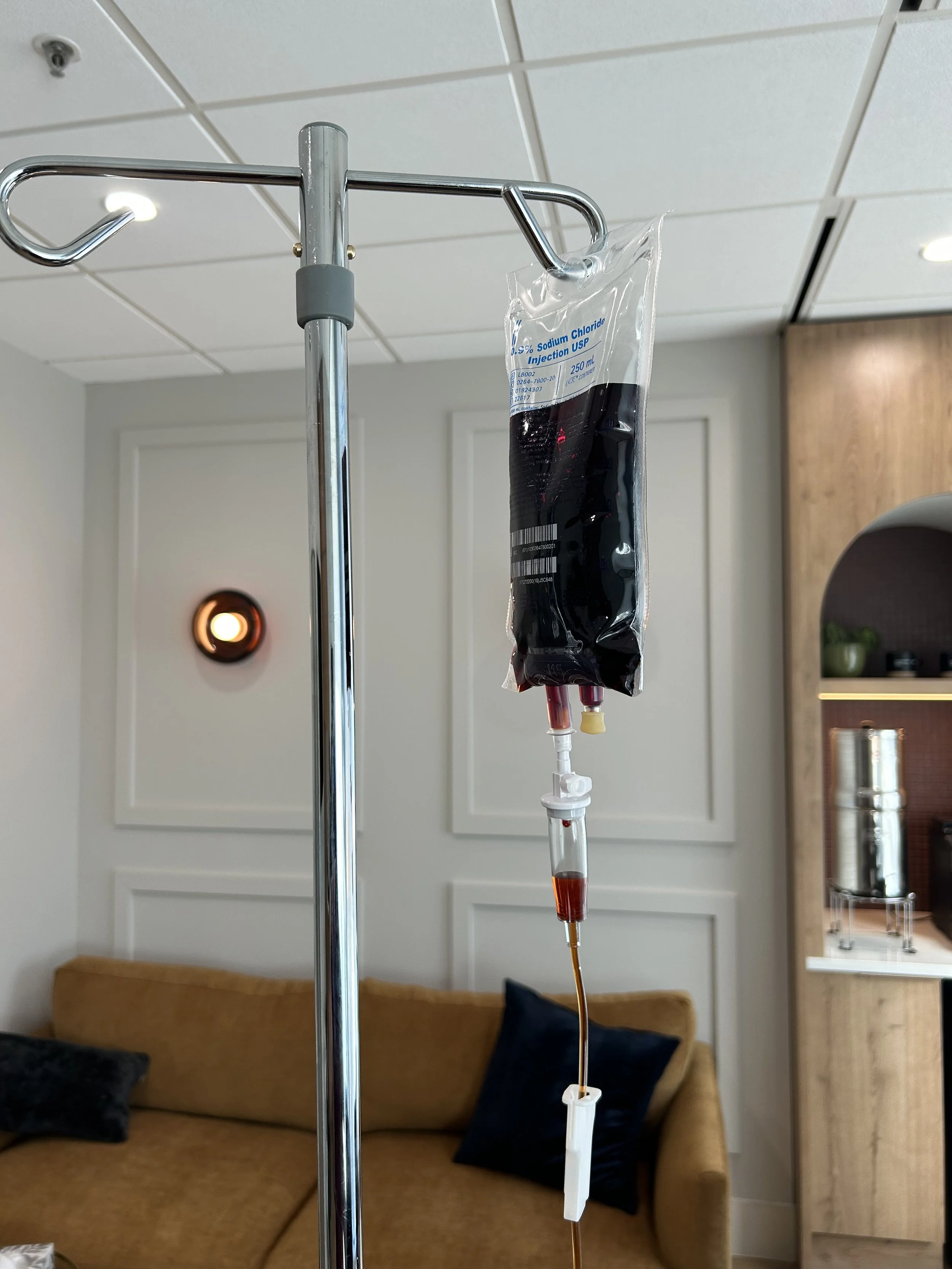 An IV drip bag filled with dark red liquid hanging from a metallic IV pole in a room with beige walls, a brown couch, and black pillows.