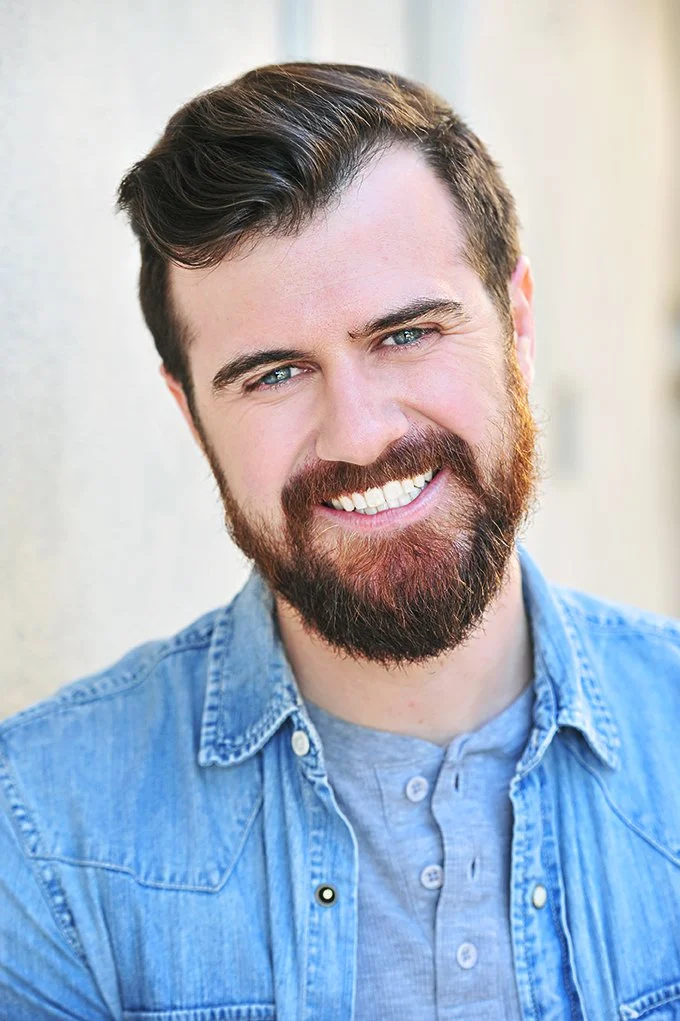 Close-up portrait of a smiling man with brown hair, blue eyes, and a beard, wearing a denim shirt over a gray T-shirt.