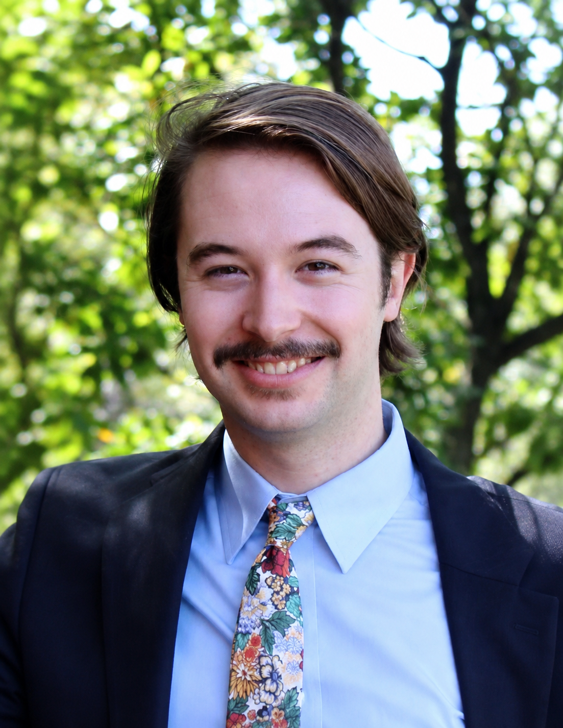 A man with brown hair, mustache, and light skin smiling outdoors, wearing a black suit jacket, light blue shirt, and a colorful floral tie, with green trees in the background.