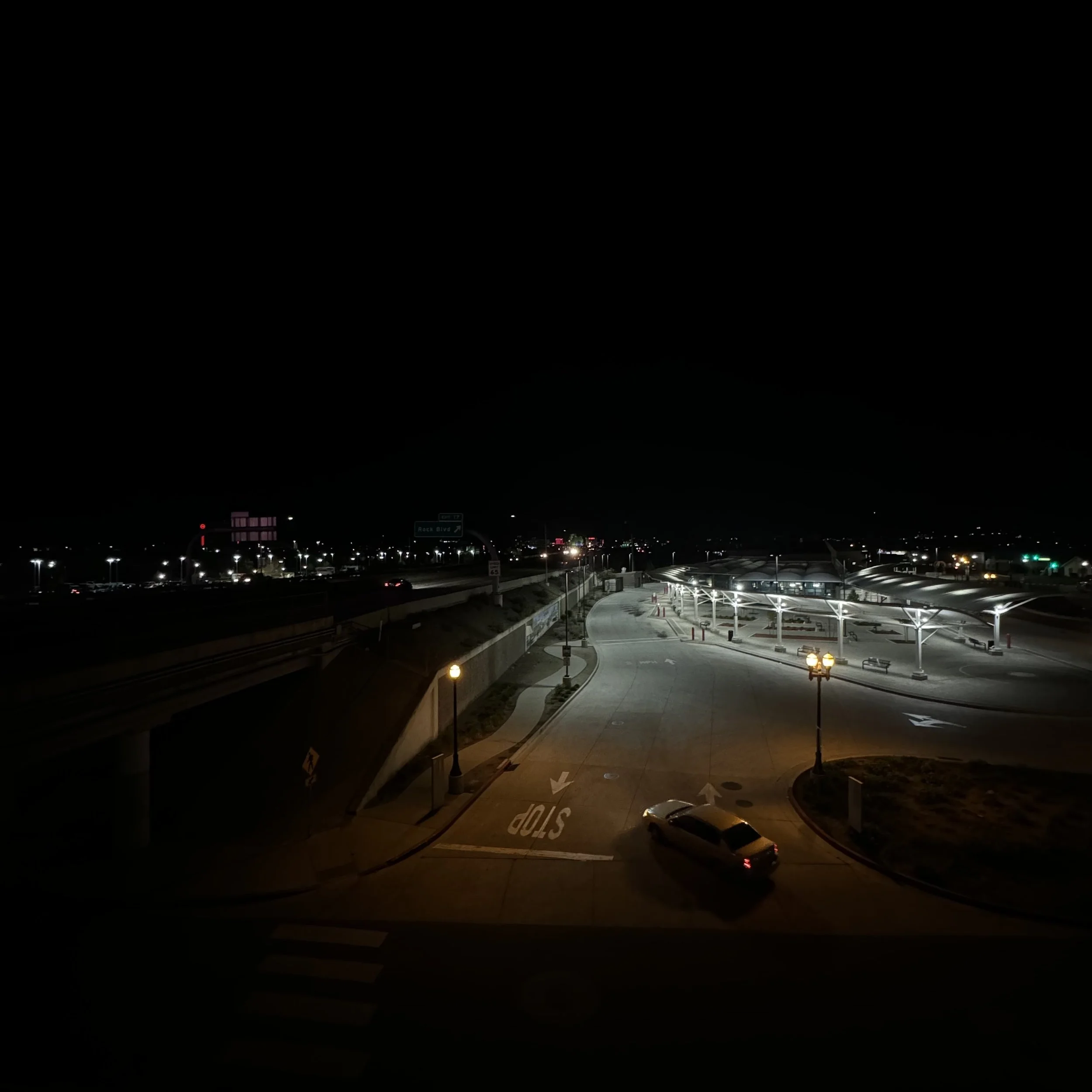 Nighttime view of a highway with cars and streetlights, a parking lot, and a building with a walkway.