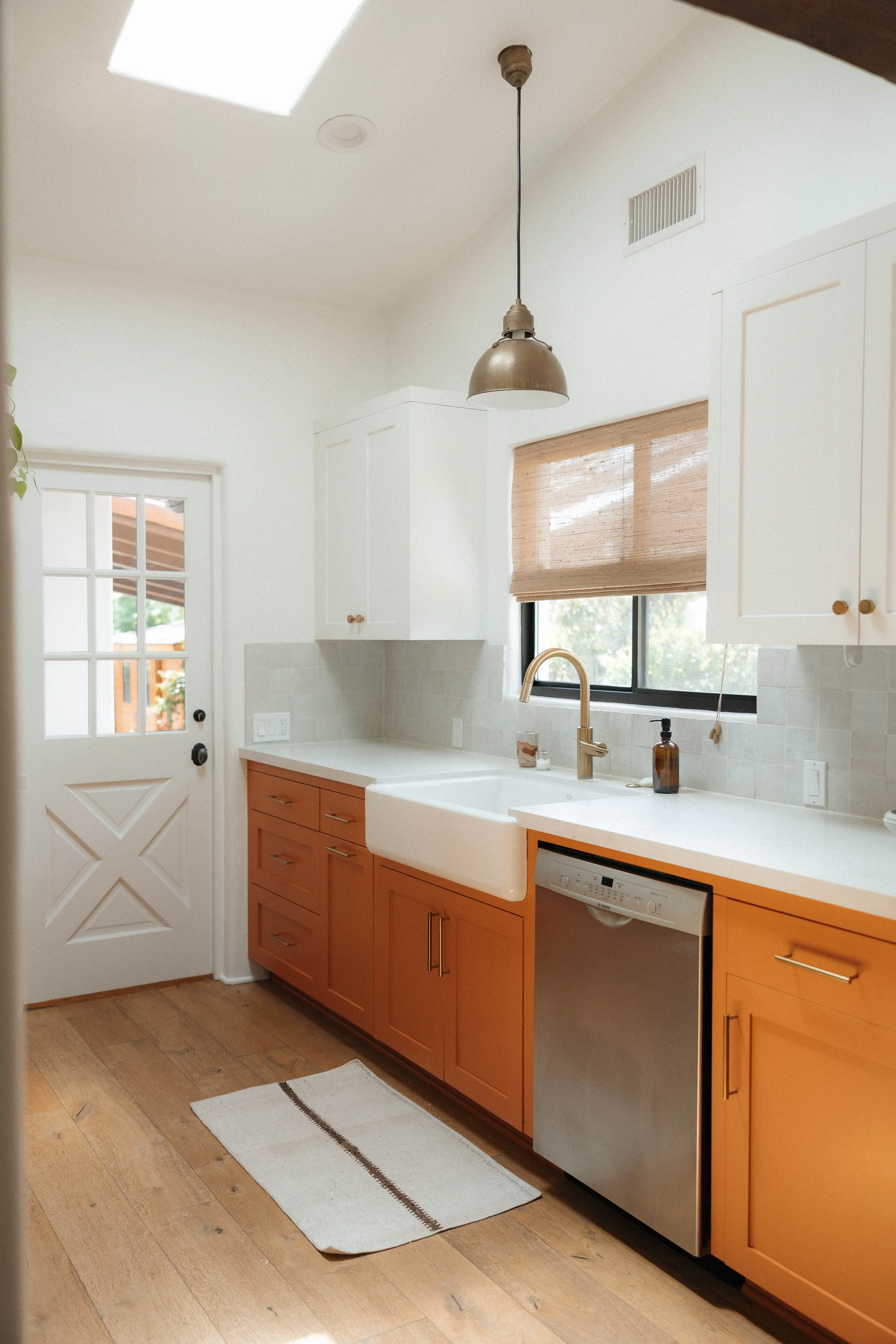A modern kitchen featuring white upper cabinets, orange lower cabinets, a white farmhouse sink with a gold faucet, and a stainless steel dishwasher. There is a window with a woven wooden shade, a white door with glass panes, a suspended pendant light