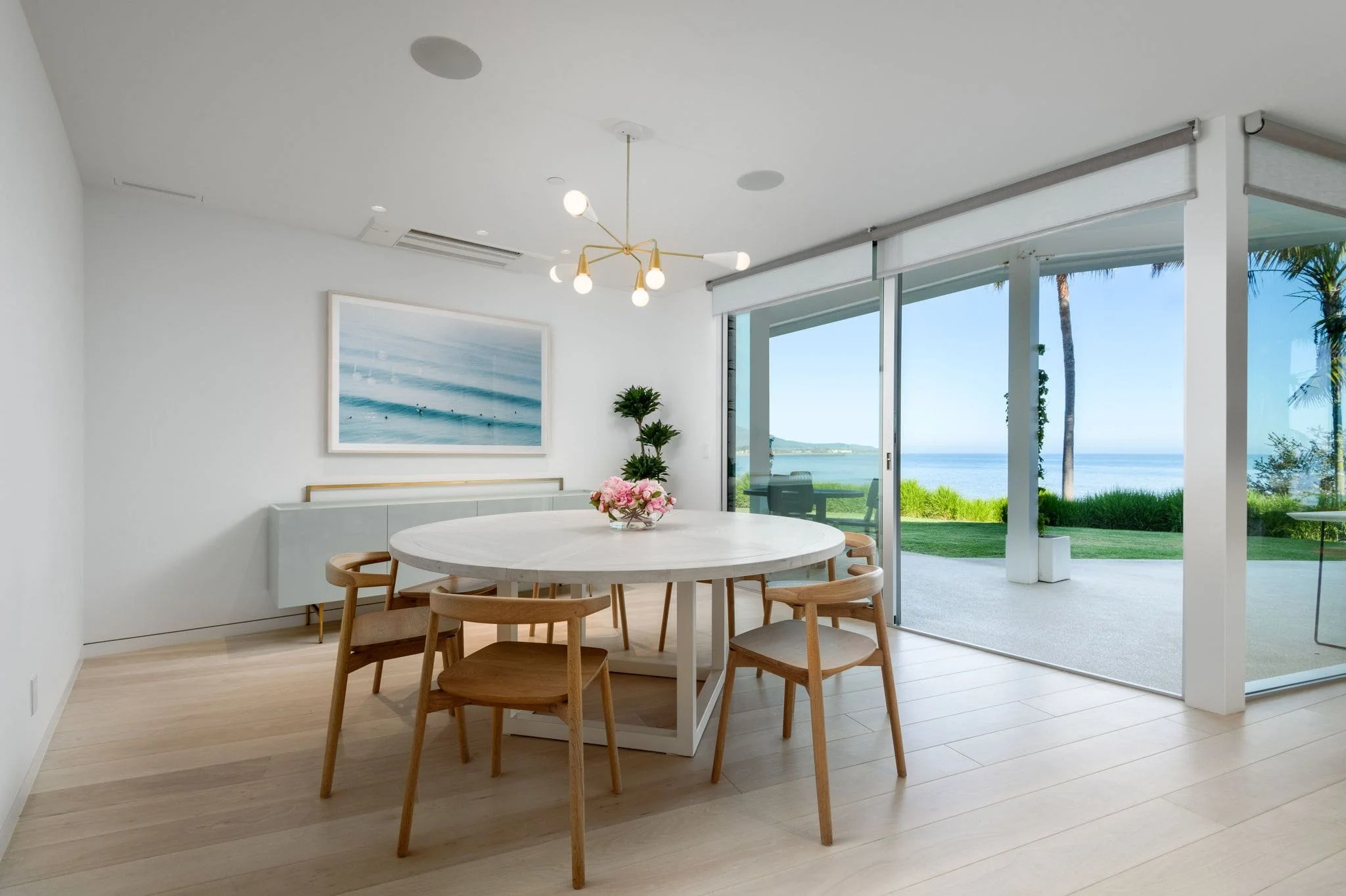 A modern, minimalist dining area with a round white table and wooden chairs, featuring a view of tropical scenery through sliding glass doors.