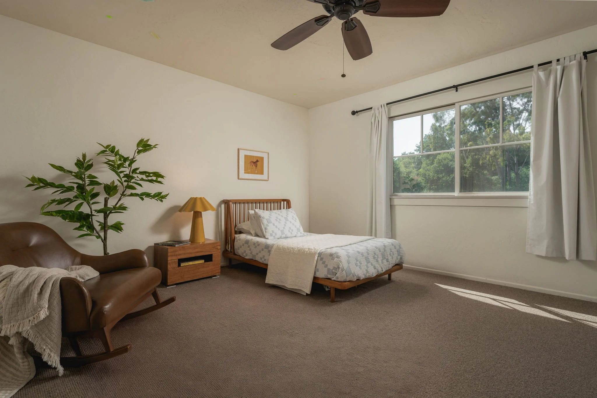 A bedroom with a bed, a wooden nightstand, a table lamp, a framed picture of a horse on the wall, a large window with white curtains, a green plant, a leather armchair with a throw blanket, a ceiling fan, and a view of trees outside.