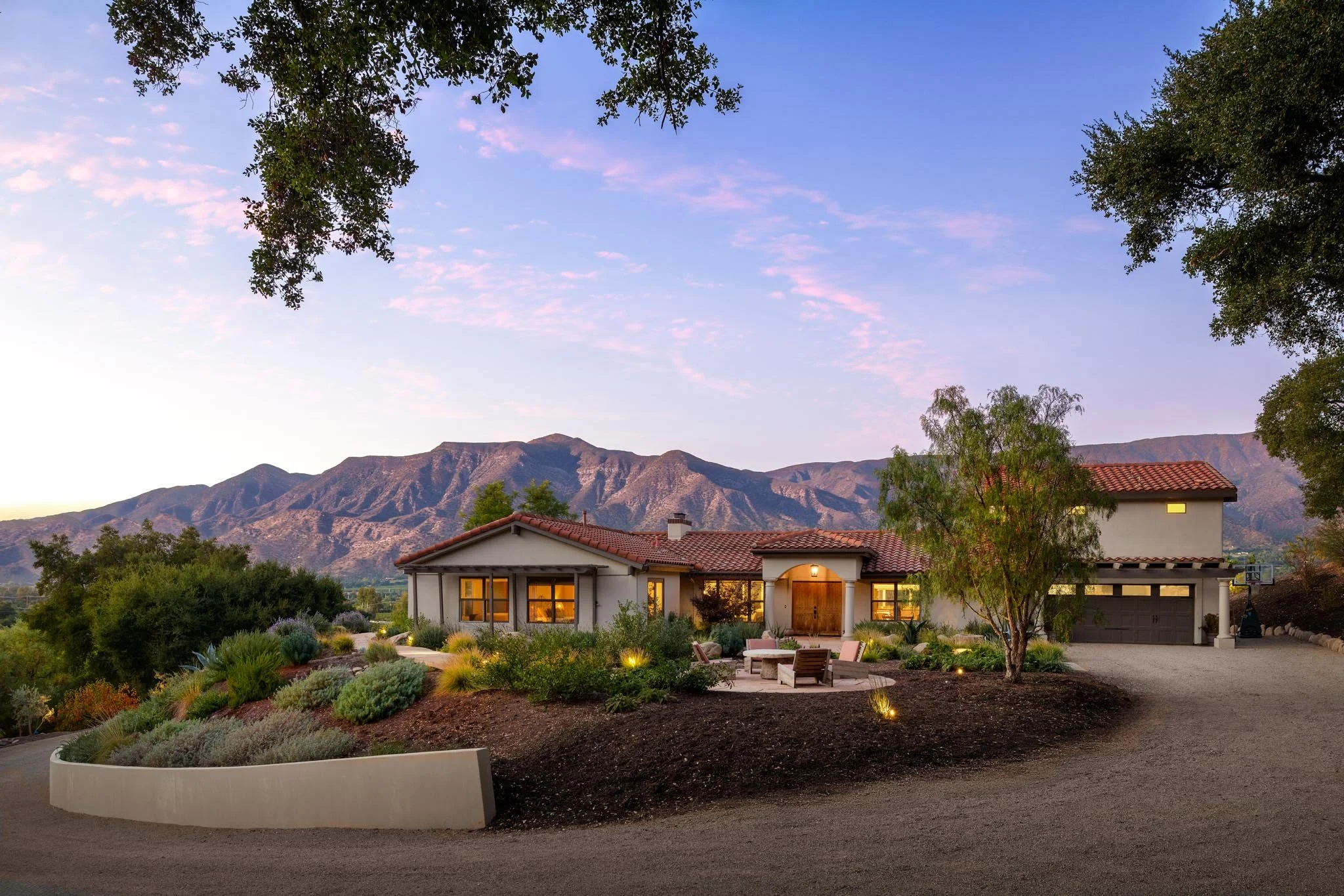 A house with a red tile roof and large windows is shown in a picturesque landscape at dusk. The house is surrounded by a well-maintained garden with various plants, trees, and outdoor furniture. Mountains are visible in the background under a sky with light clouds, and the scene is illuminated with warm outdoor lighting.