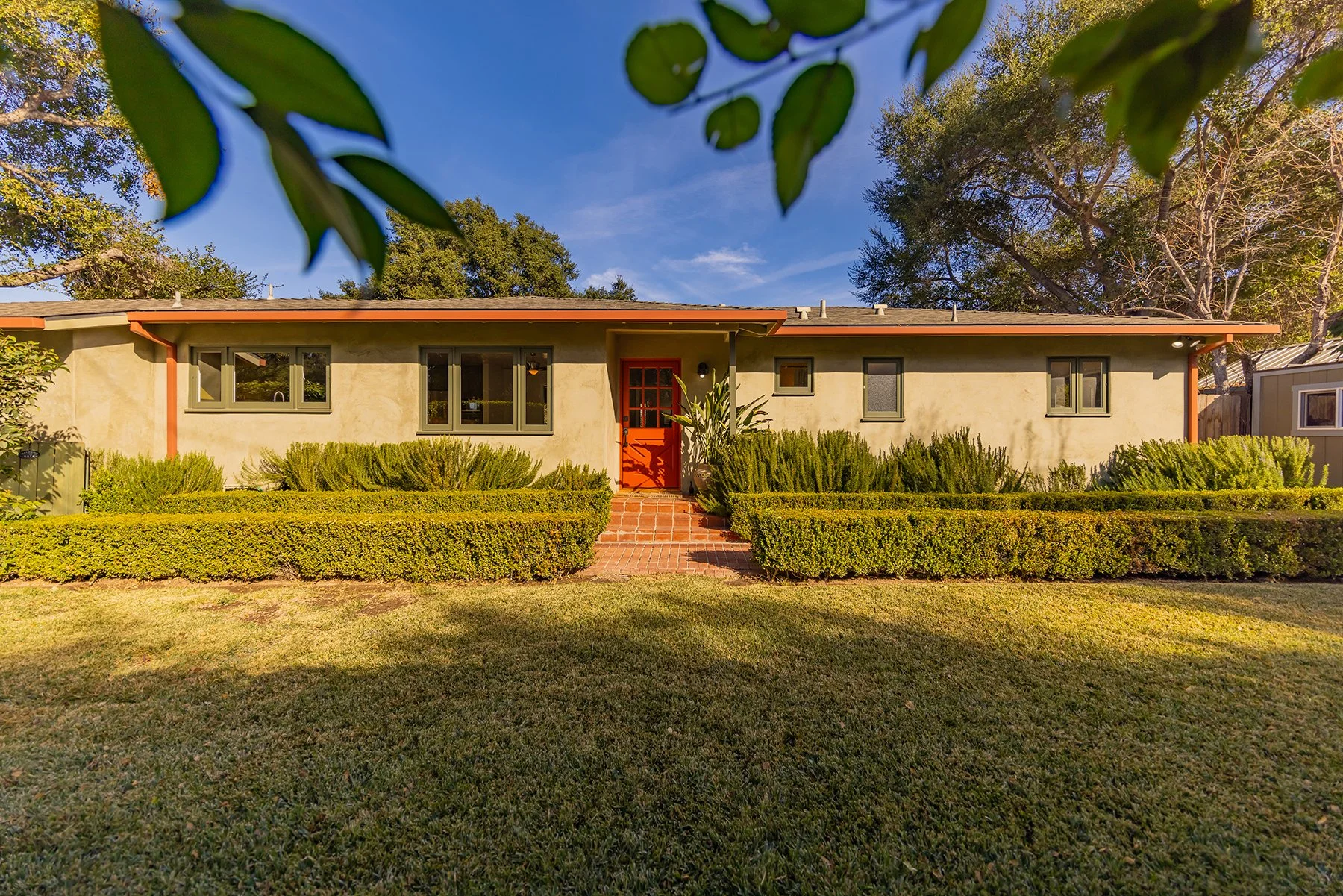 Front of a single-story house with a red door, surrounded by green bushes and plants, with a brick walkway and a lawn, under a clear blue sky.