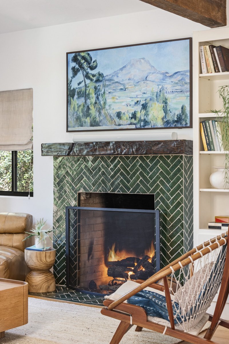 Living room with a fireplace, green herringbone tile surround, painting of a mountain landscape above, wood mantel, bookshelf with books, and furniture including a leather chair, a wooden side table with a plant, and a wooden chair with a patterned c