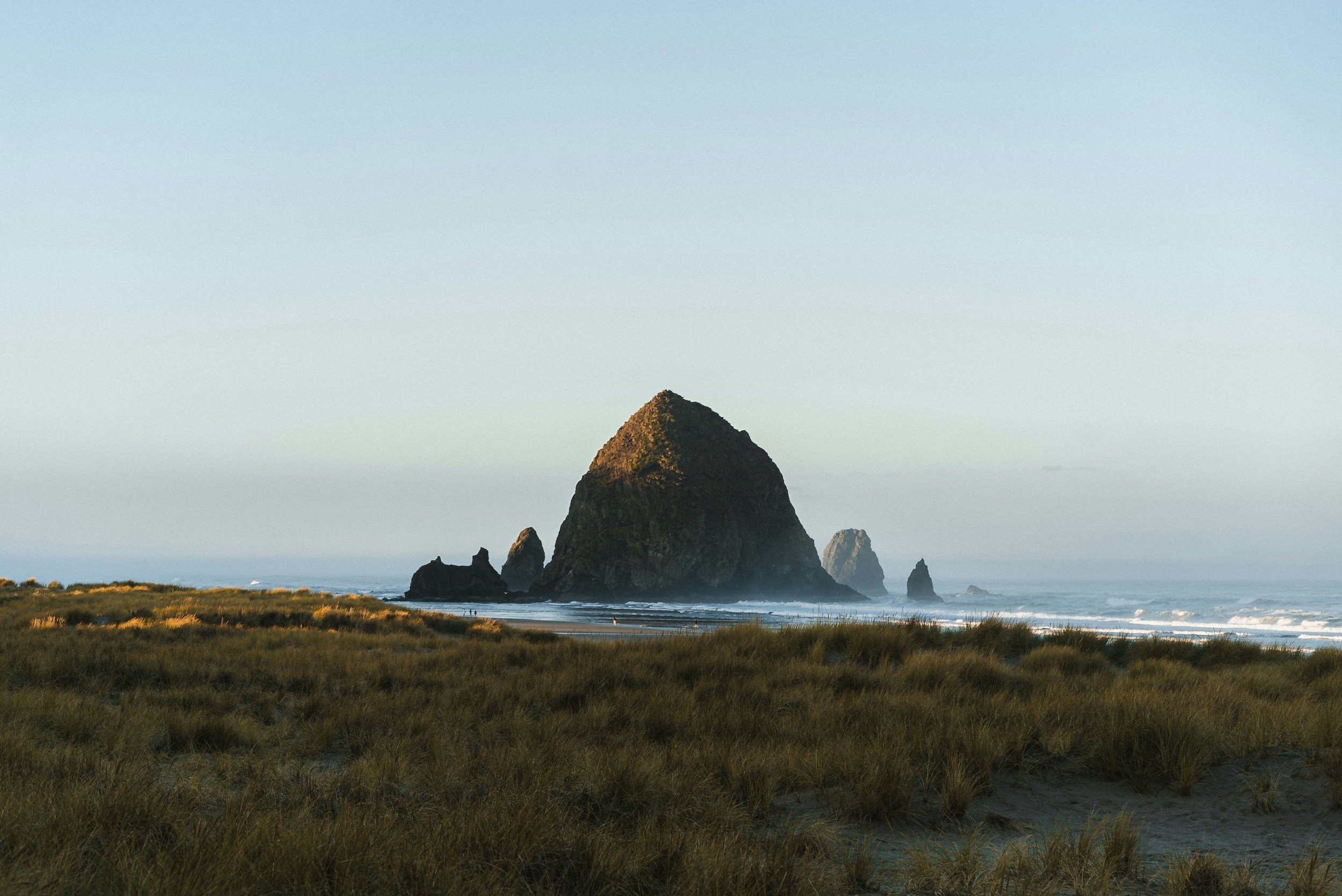 A large rock formation rises from the ocean near a grassy shoreline under a clear sky.
