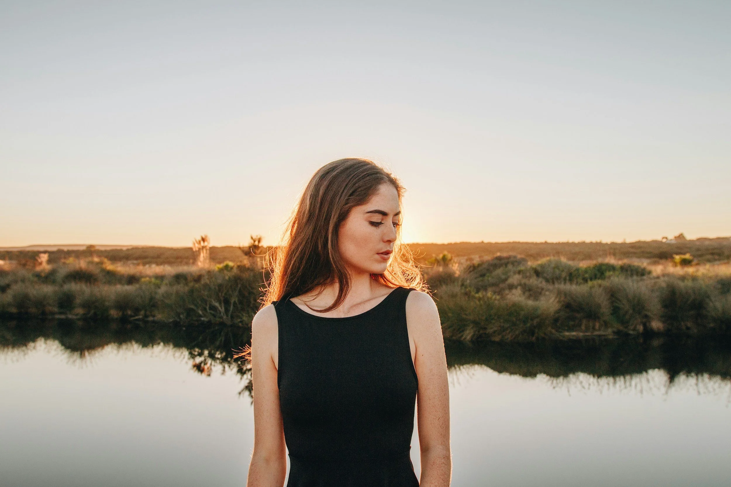 A young woman with long brown hair in a black sleeveless top standing outdoors near a body of water during sunset, with trees in the background.