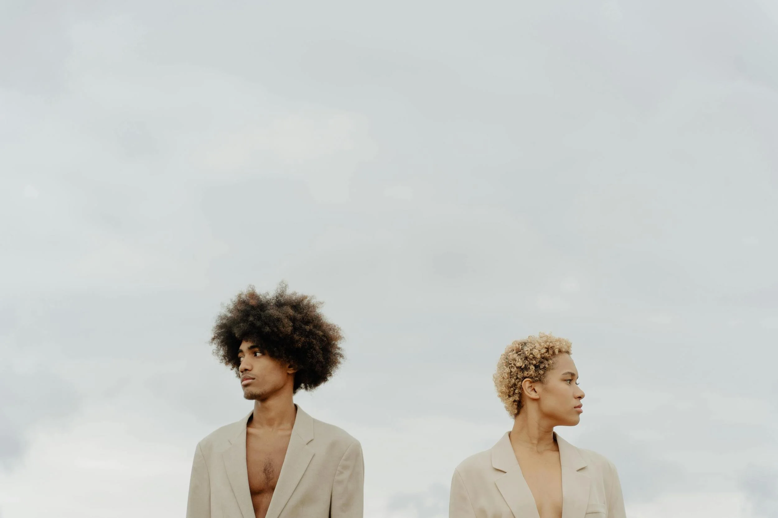 Two individuals, one man with dark curly hair and a woman with blonde curly hair, standing outdoors against a cloudy sky, both wearing beige blazers, facing opposite directions.