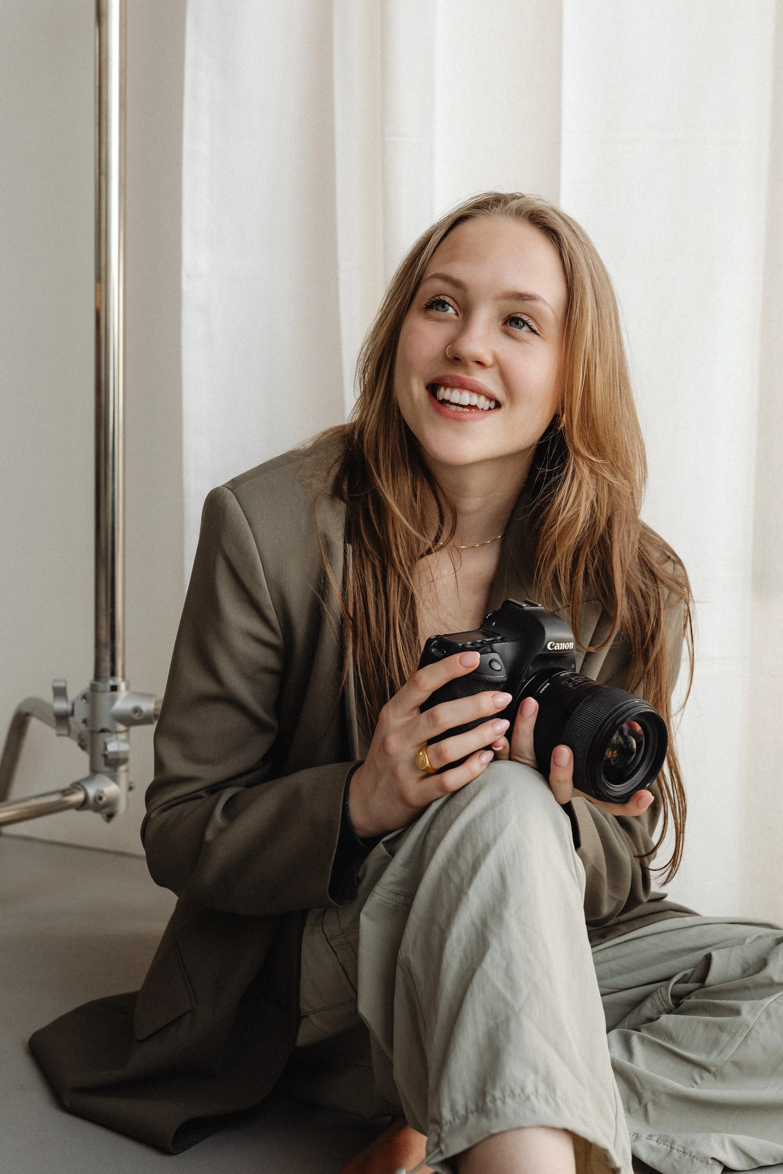 A young woman with long red hair and a nose ring is sitting on the floor, smiling, holding a professional camera in her hands. She is dressed in a khaki blazer and light-colored pants, with a relaxed, casual pose near a white wall with natural light.