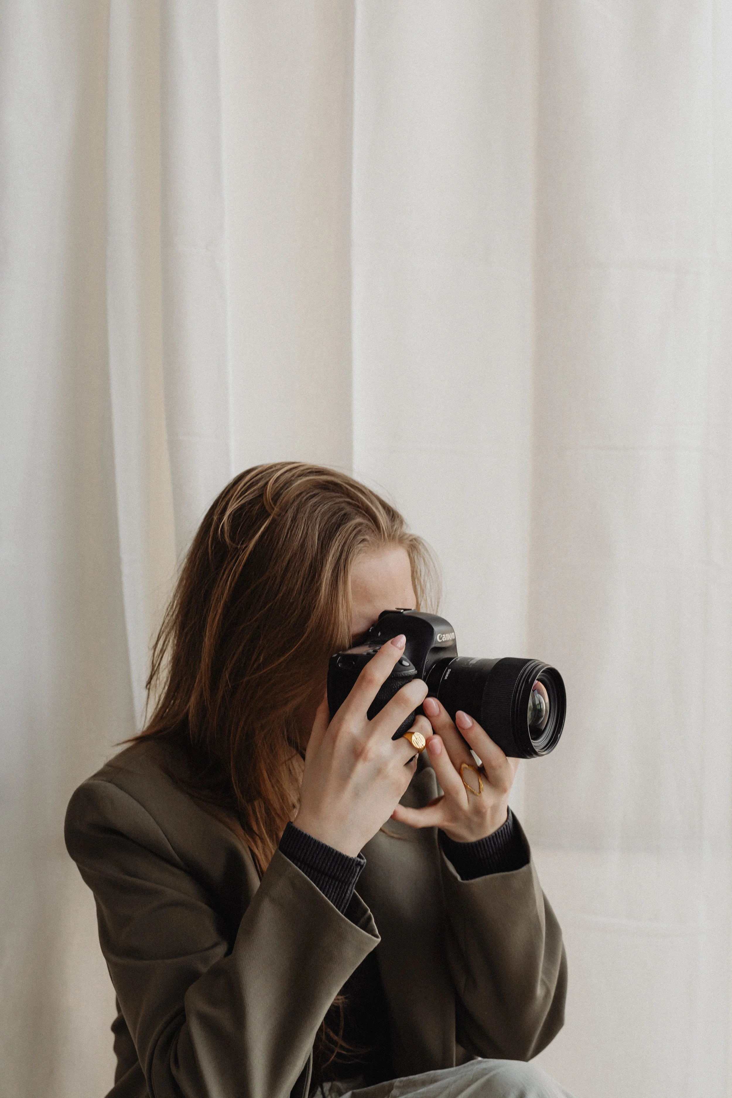 A woman with long, light brown hair taking a photograph with a black camera, wearing a brown jacket and gold rings, sitting in front of light-colored curtains.