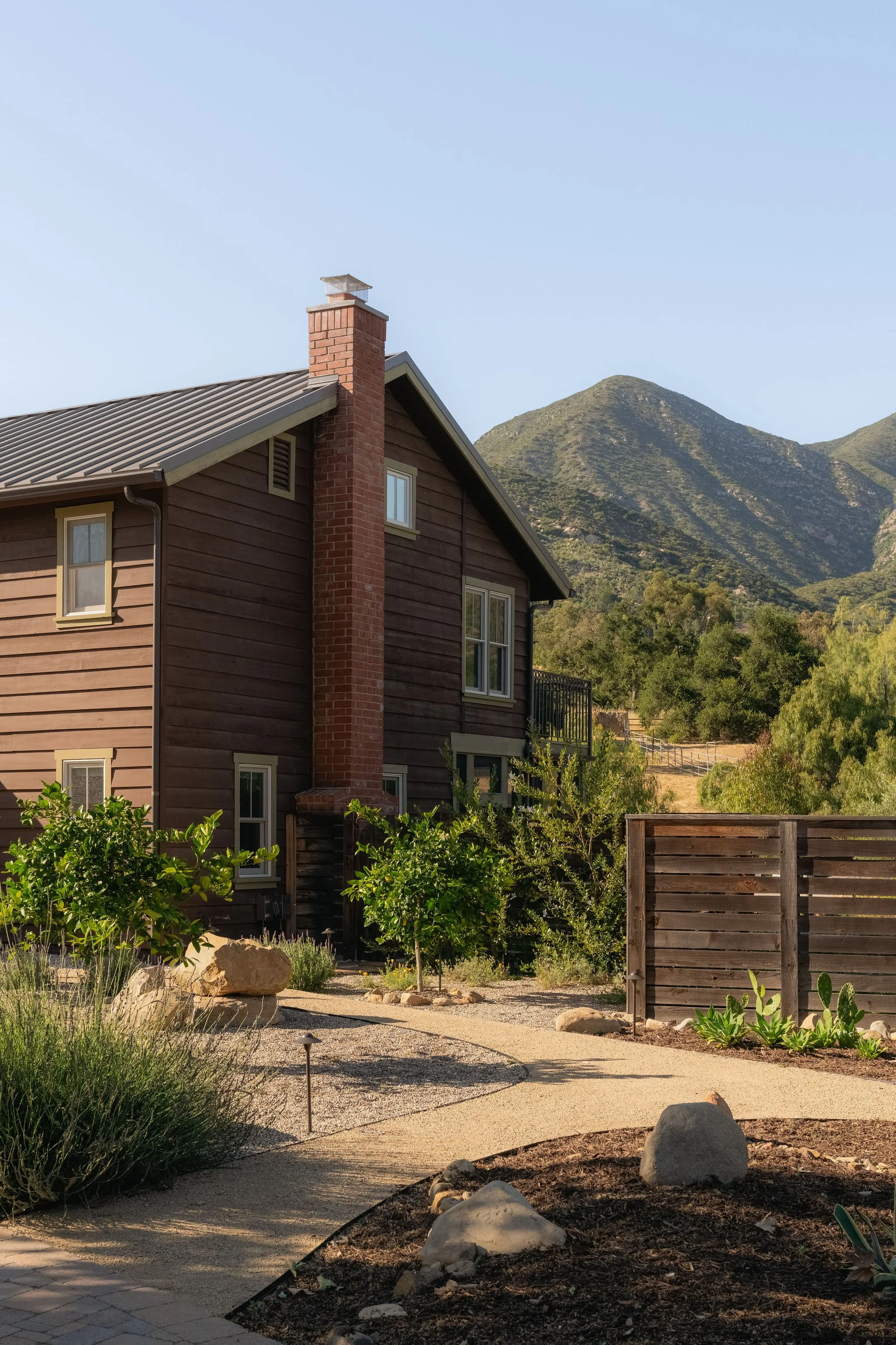 House with brown wooden siding, brick chimney, and large windows, surrounded by a landscaped yard with bushes, rocks, and a gravel path, with mountains in the background and a clear sky.