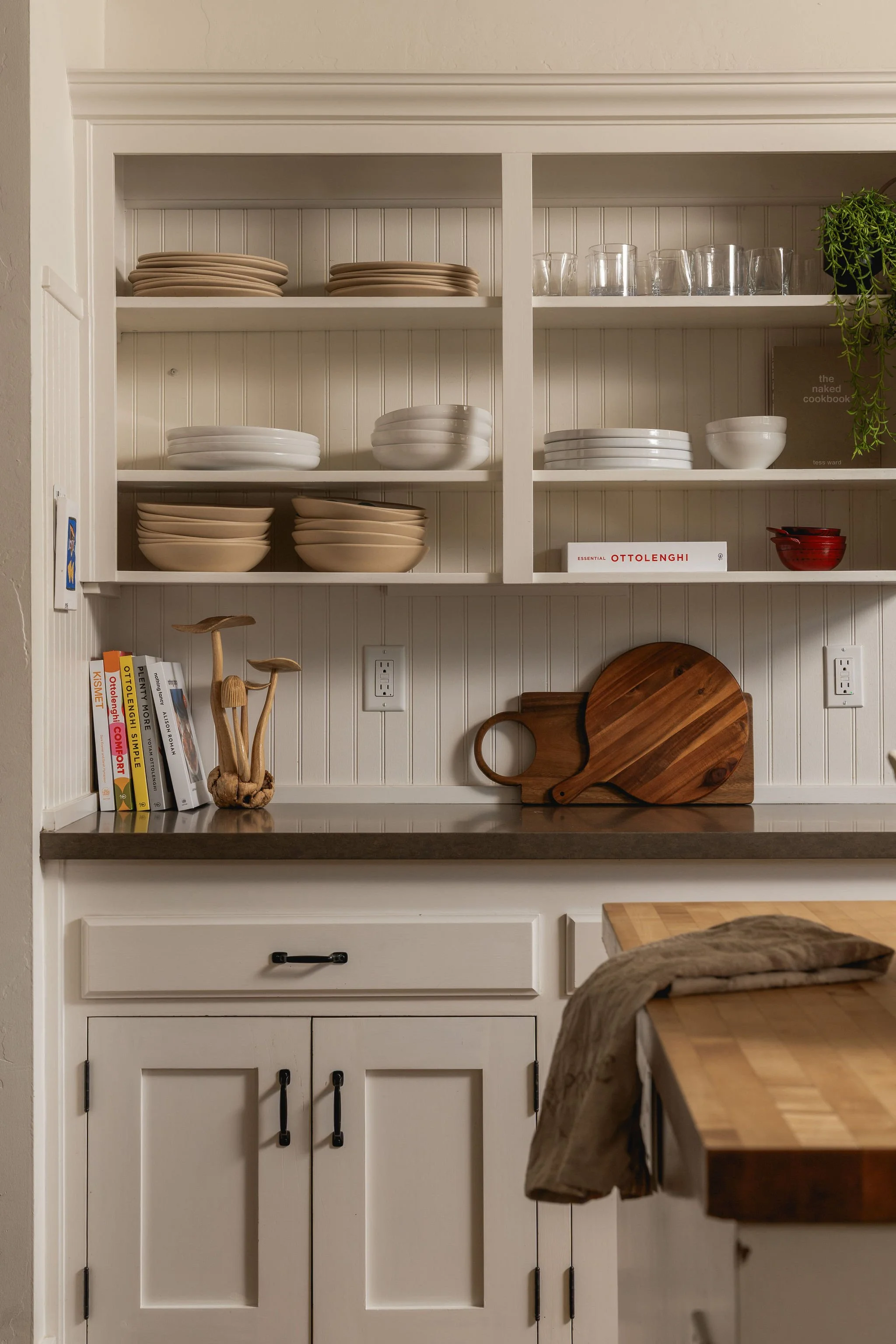 Open white kitchen cabinet filled with white and beige dishes and glasses, a black book titled 'the naked cookbook,' a green plant, a round wooden cutting board, and a wooden spoon holder on a dark countertop, with a wooden cutting board and cloth on