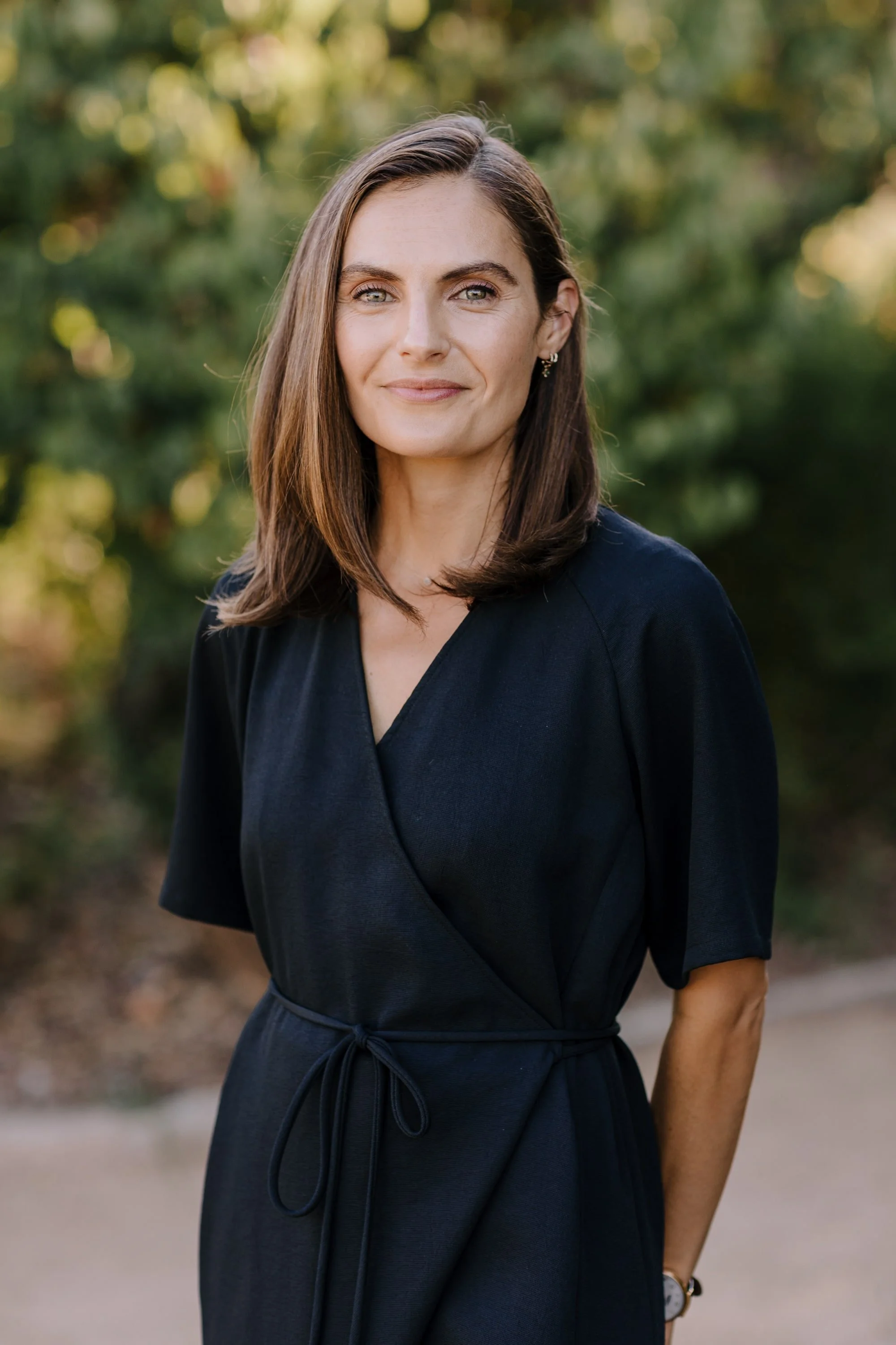 A woman with shoulder-length brown hair, wearing a black dress and earrings, standing outdoors with blurred green trees and sunlight in the background.
