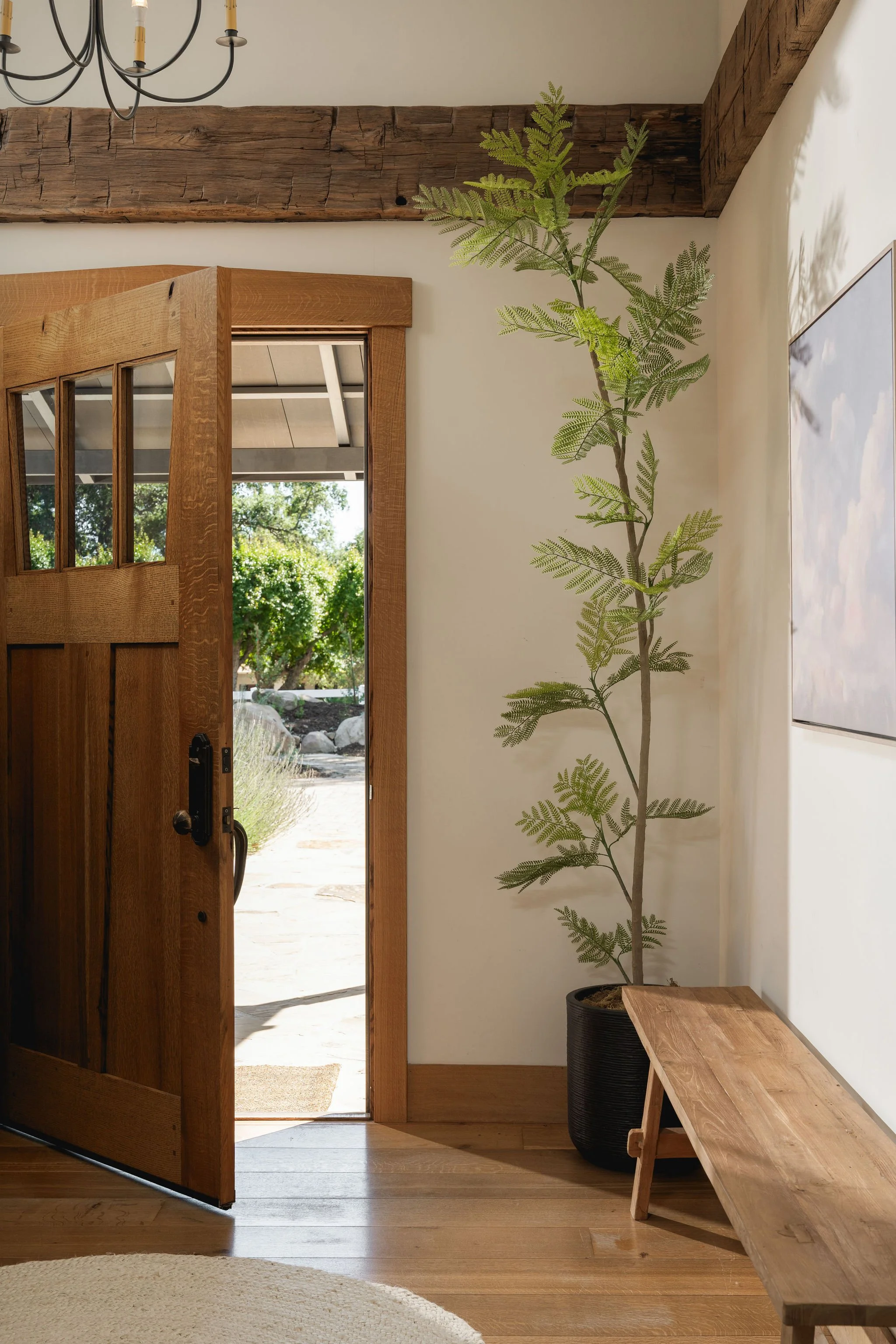 Open wooden door leading outside, with a view of trees and rocks, inside a room with light wood flooring, a potted plant in a black container, and a wooden bench. An exposed wooden beam and a chandelier are visible on the ceiling.