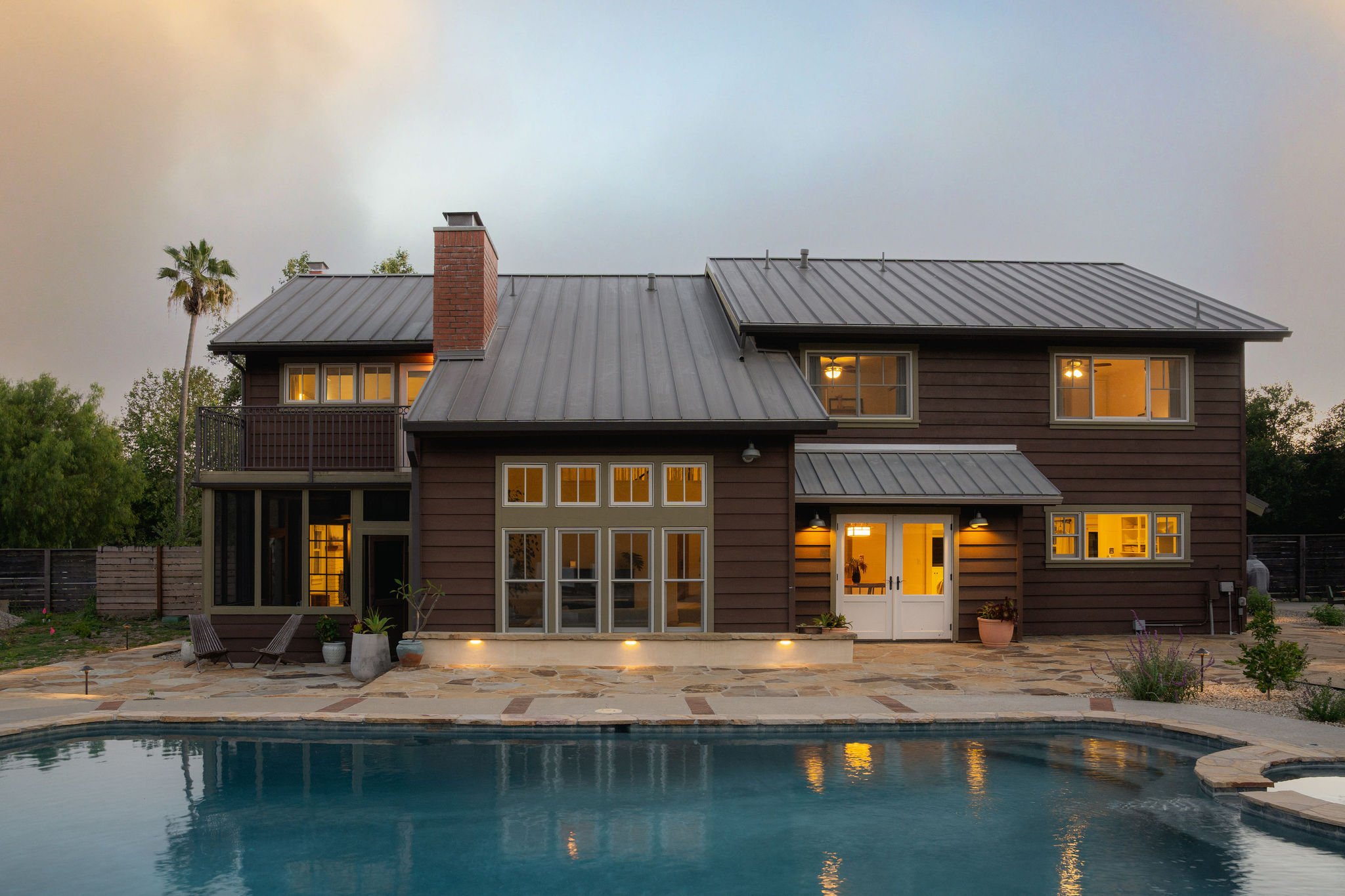 Backyard scene featuring a two-story house with large windows, a metal roof, a brick chimney, and a backyard with a swimming pool, patio furniture, potted plants, and surrounding greenery at dusk.